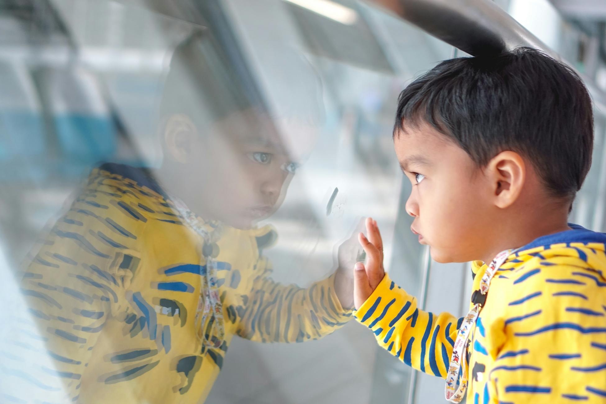 A cute child thoughtfully gazing at his reflection on a glass wall indoors, wearing a colorful hoodie.