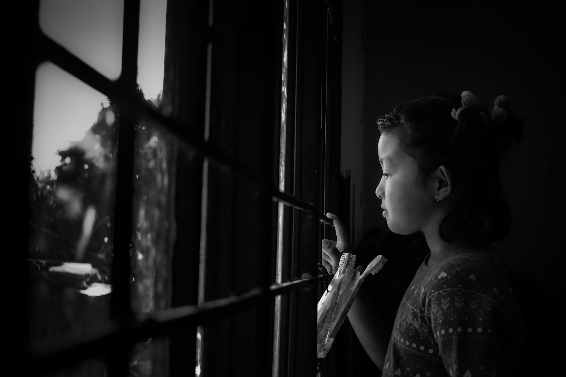 Side view of black and white adorable pensive little Asian girl standing in bathroom and looking out fenced window on rainy day