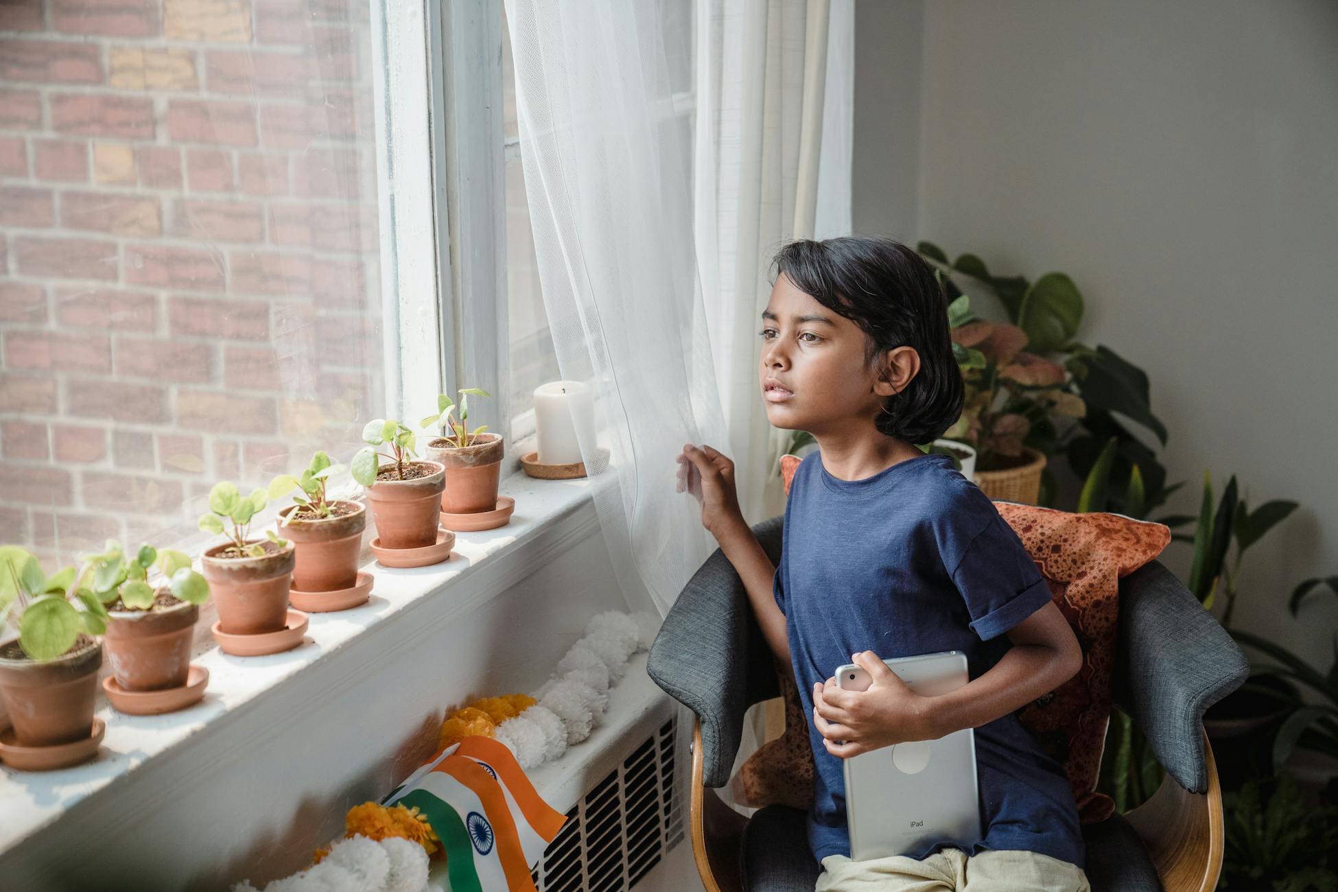 Young boy sitting indoors, gazing out the window, holding a tablet near Indian flag.