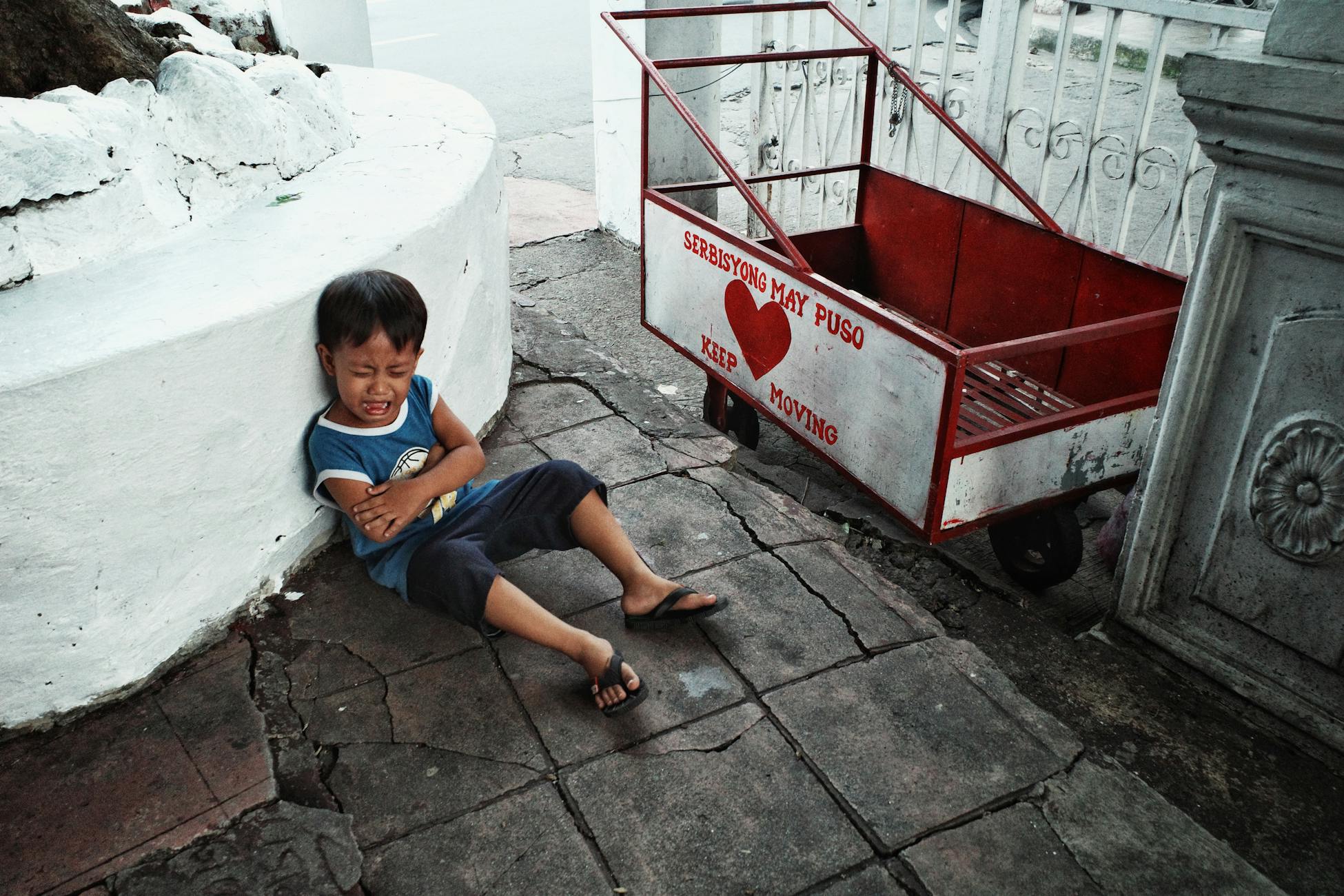 Candid street photograph of a crying child sitting on a city sidewalk near a red cart.