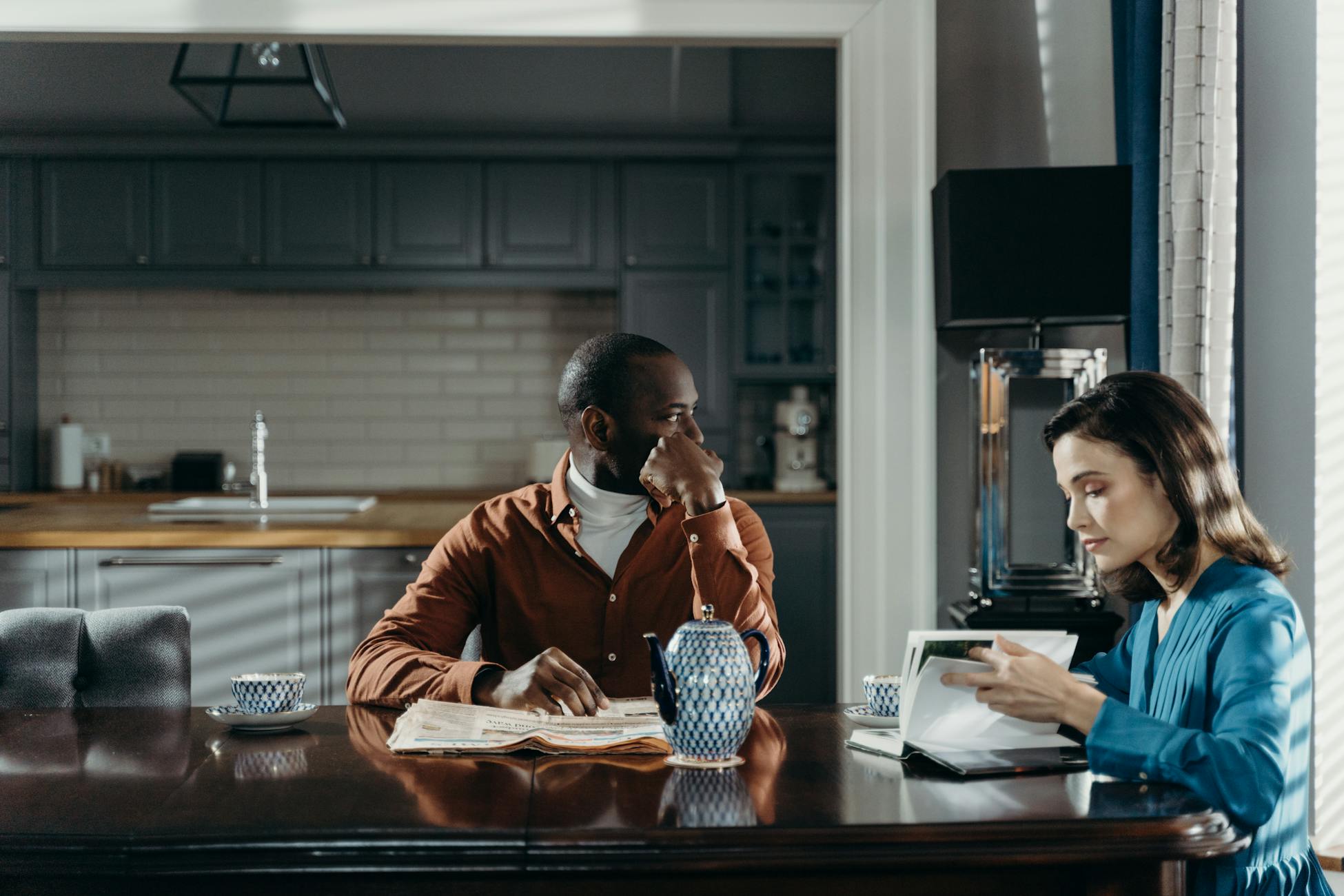 A couple sits at a dining table in a modern kitchen, reading and relaxing.
