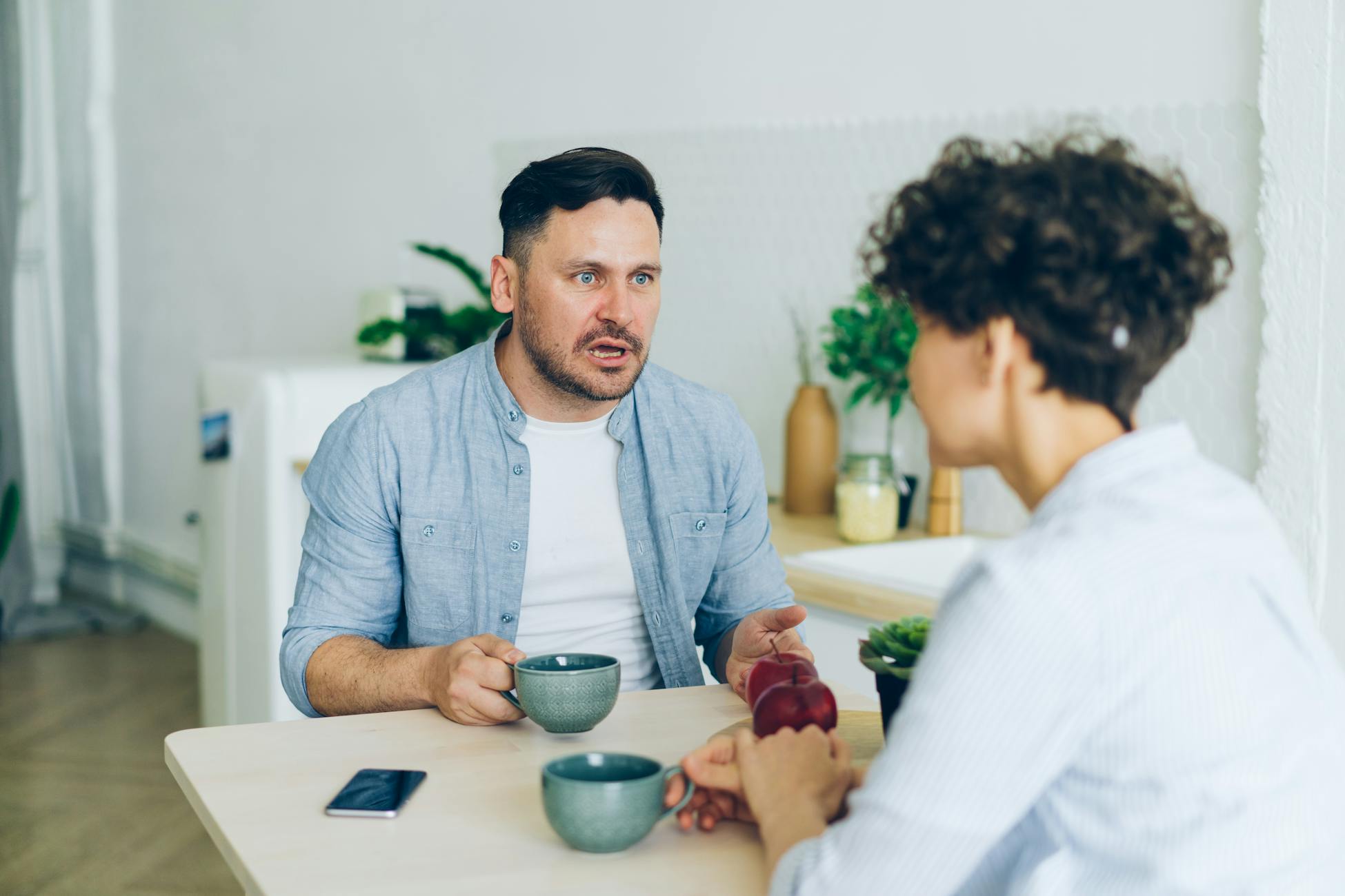 Couple having a serious conversation at the kitchen table with coffee cups.