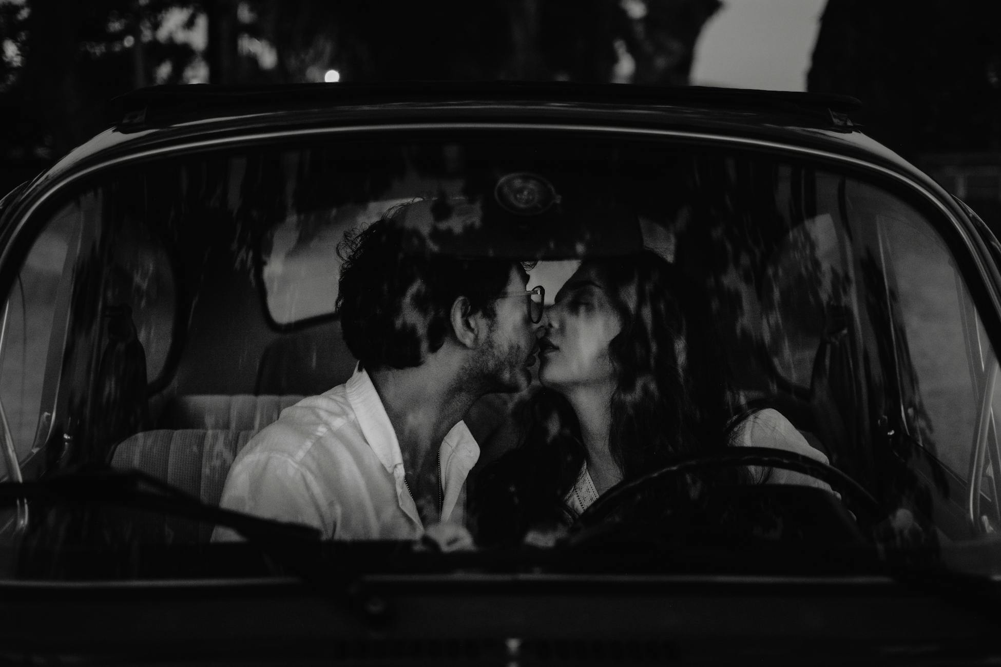 A black and white image of an affectionate couple kissing inside a vintage car.