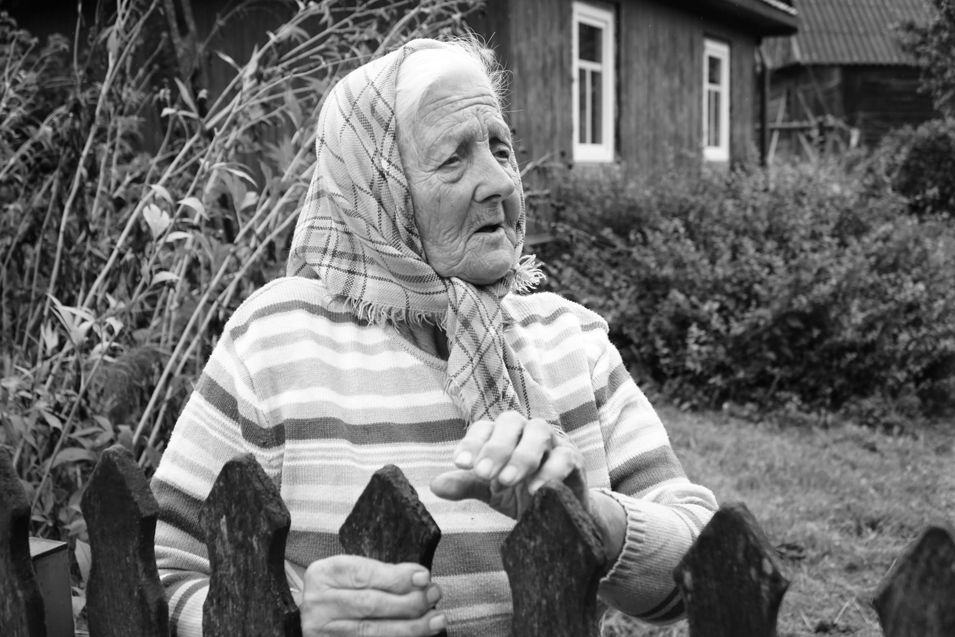 Elderly woman with headscarf outdoors near rustic wooden fence and cottage.