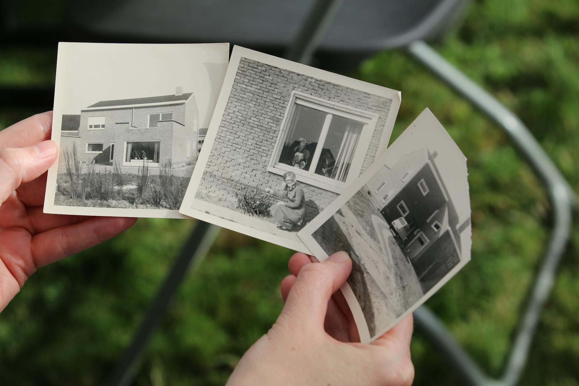 Hands holding vintage black and white photos of old houses outdoors.