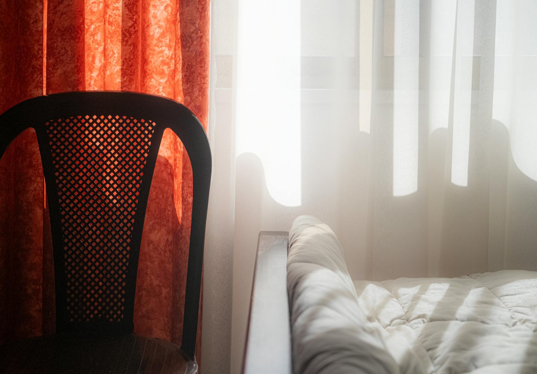 Sunlight streaming through curtains illuminating a bed and chair in a cozy bedroom setting.