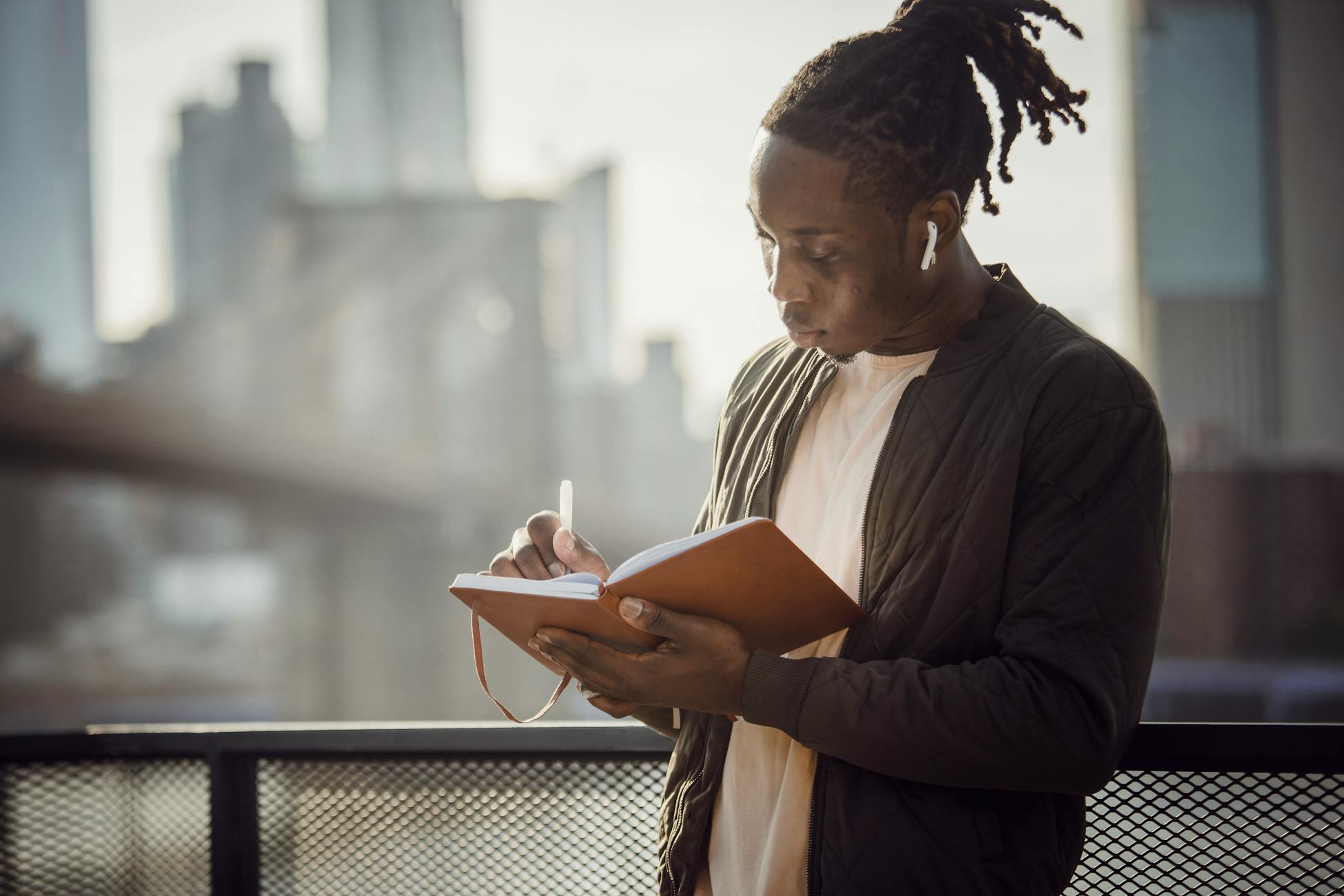 Young African American male author writing in notebook and listening to music through wireless earbuds while standing in back lit on open terrace against blurry modern city