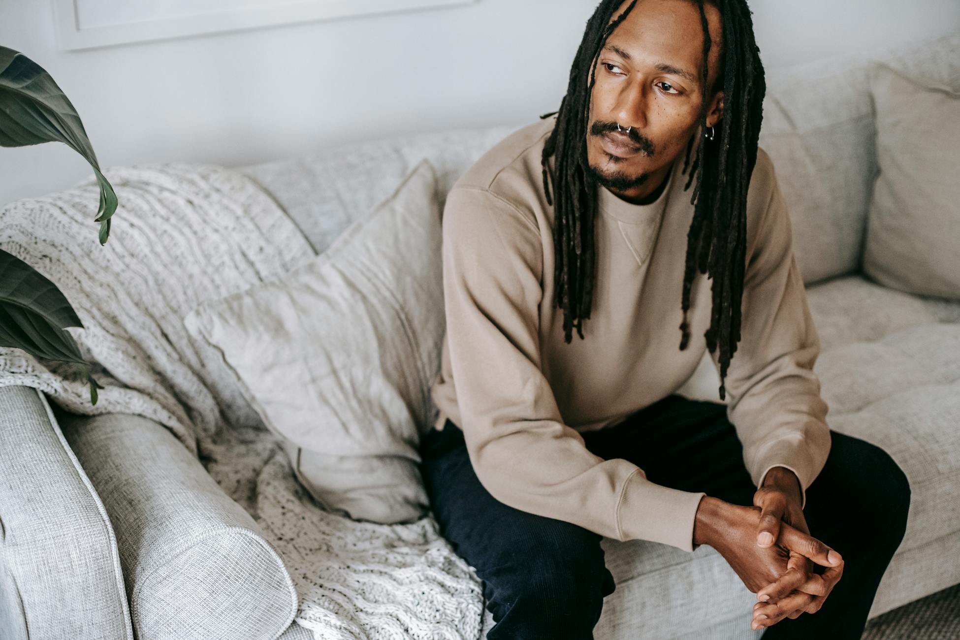 Pensive African American male in casual outfit with dreadlocks sitting on couch with folded hands and thinking while looking away