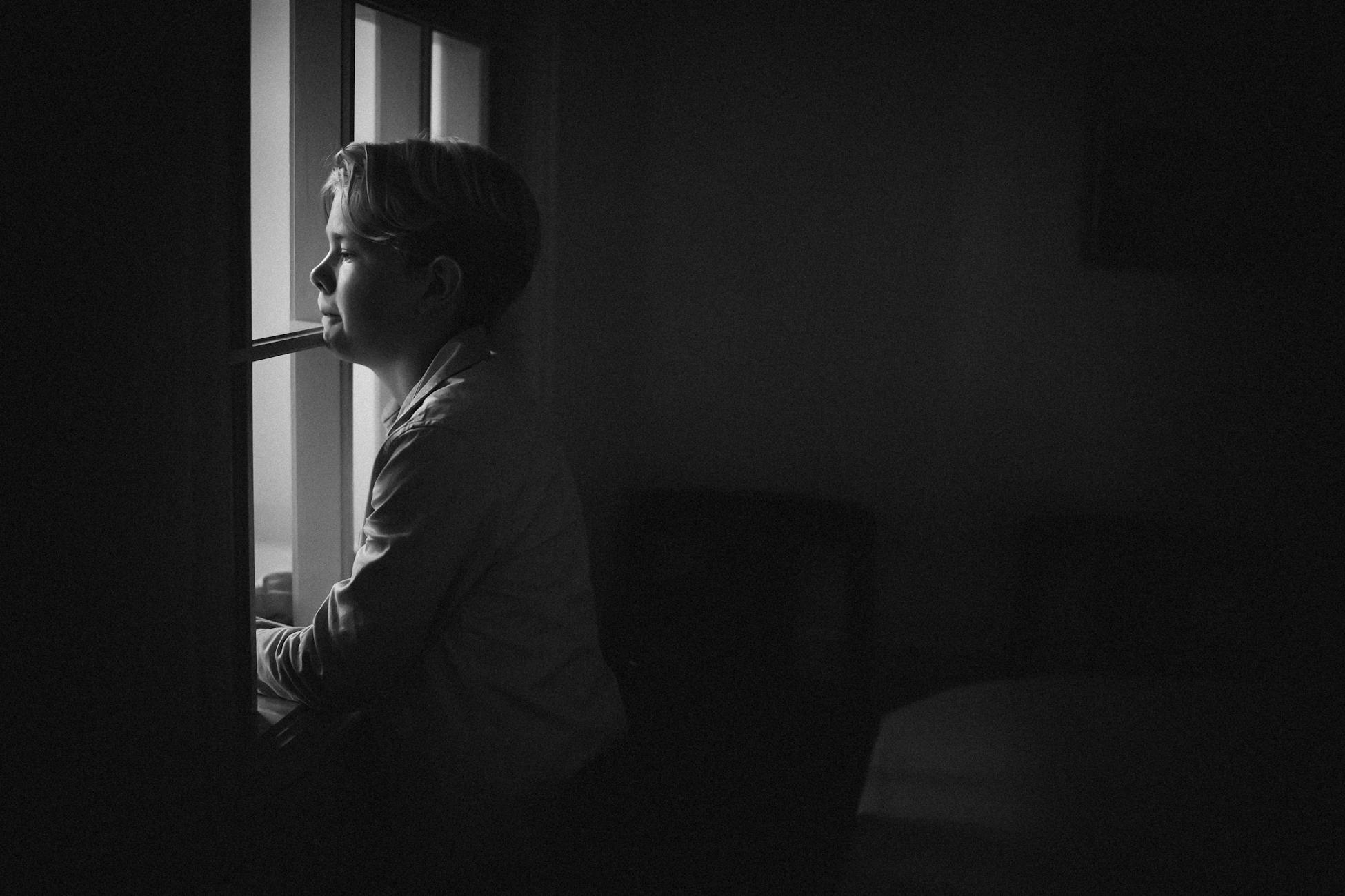 A contemplative child gazes out the window in a moody black and white scene.
