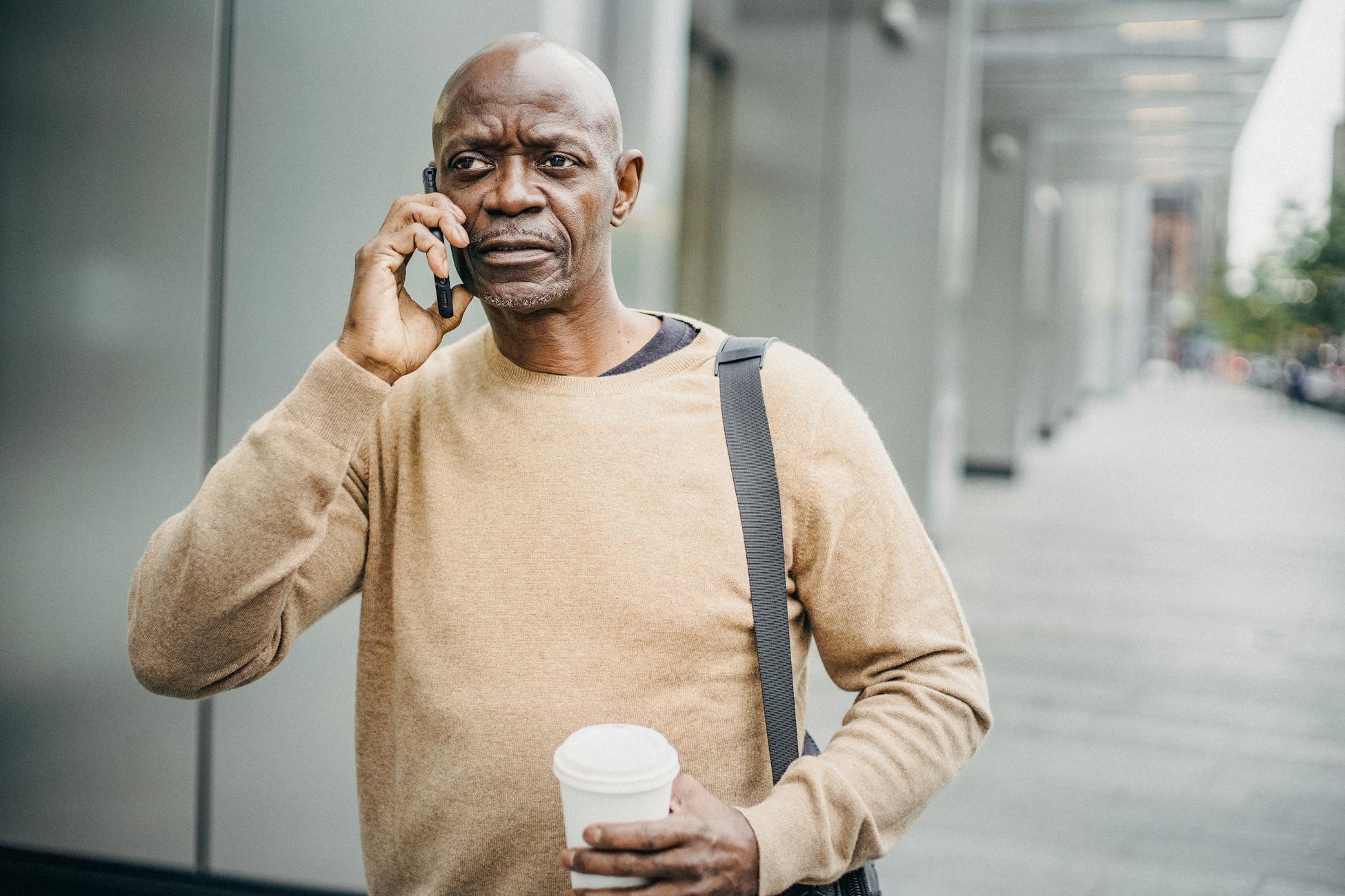 Focused African American male with coffee cup chatting on mobile phone walking down street