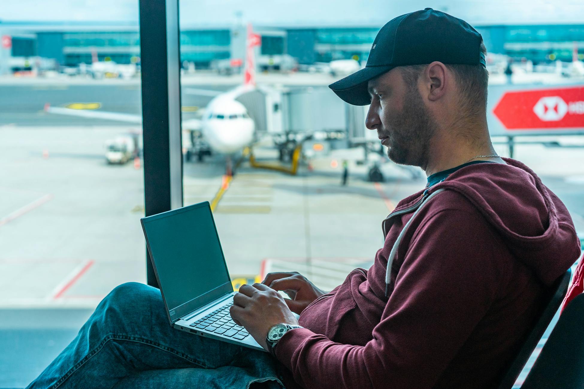 A man working on his laptop in an airport terminal, with a plane visible outside.