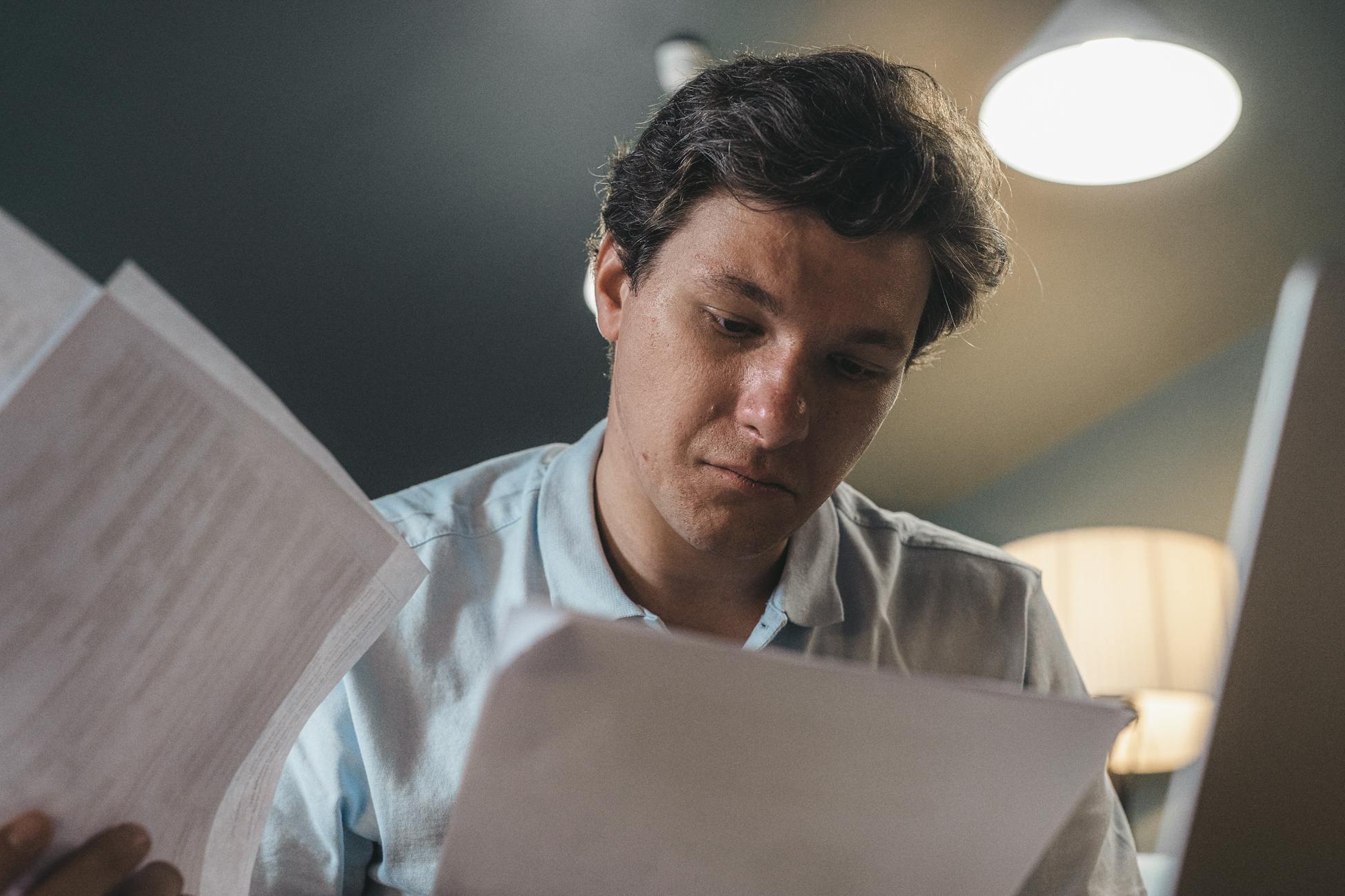 Serious man wearing a light blue shirt intently reading printed papers indoors under warm lighting.