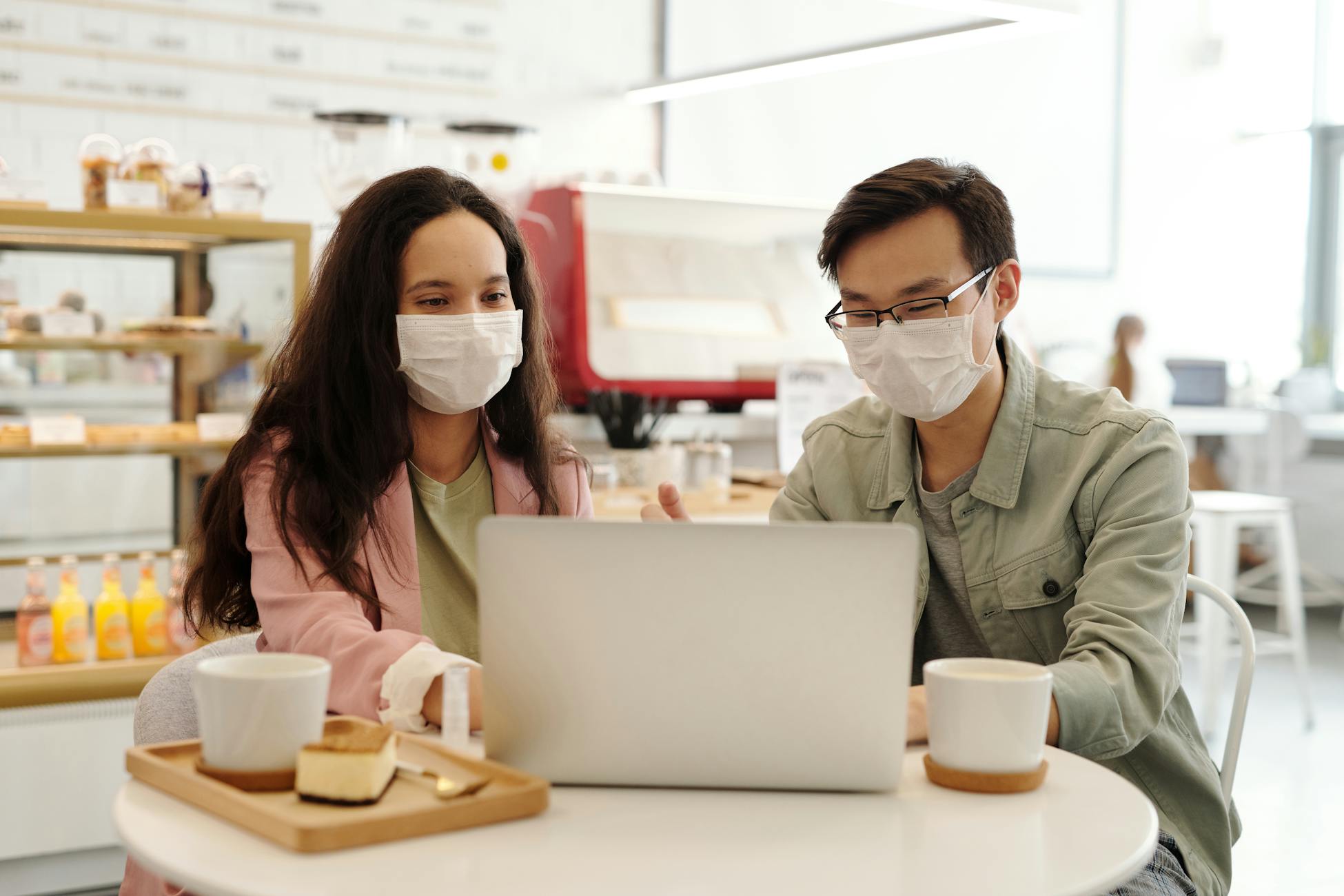 Two people wearing face masks discuss work on a laptop in a cafe setting.