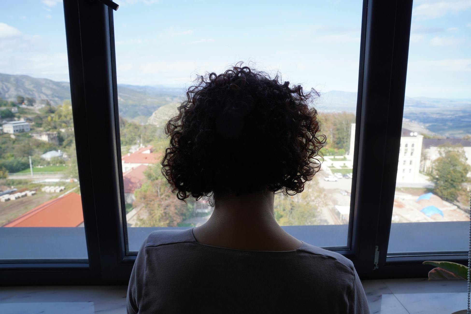 A woman with curly hair gazing out a window at a scenic mountain view, indoors.