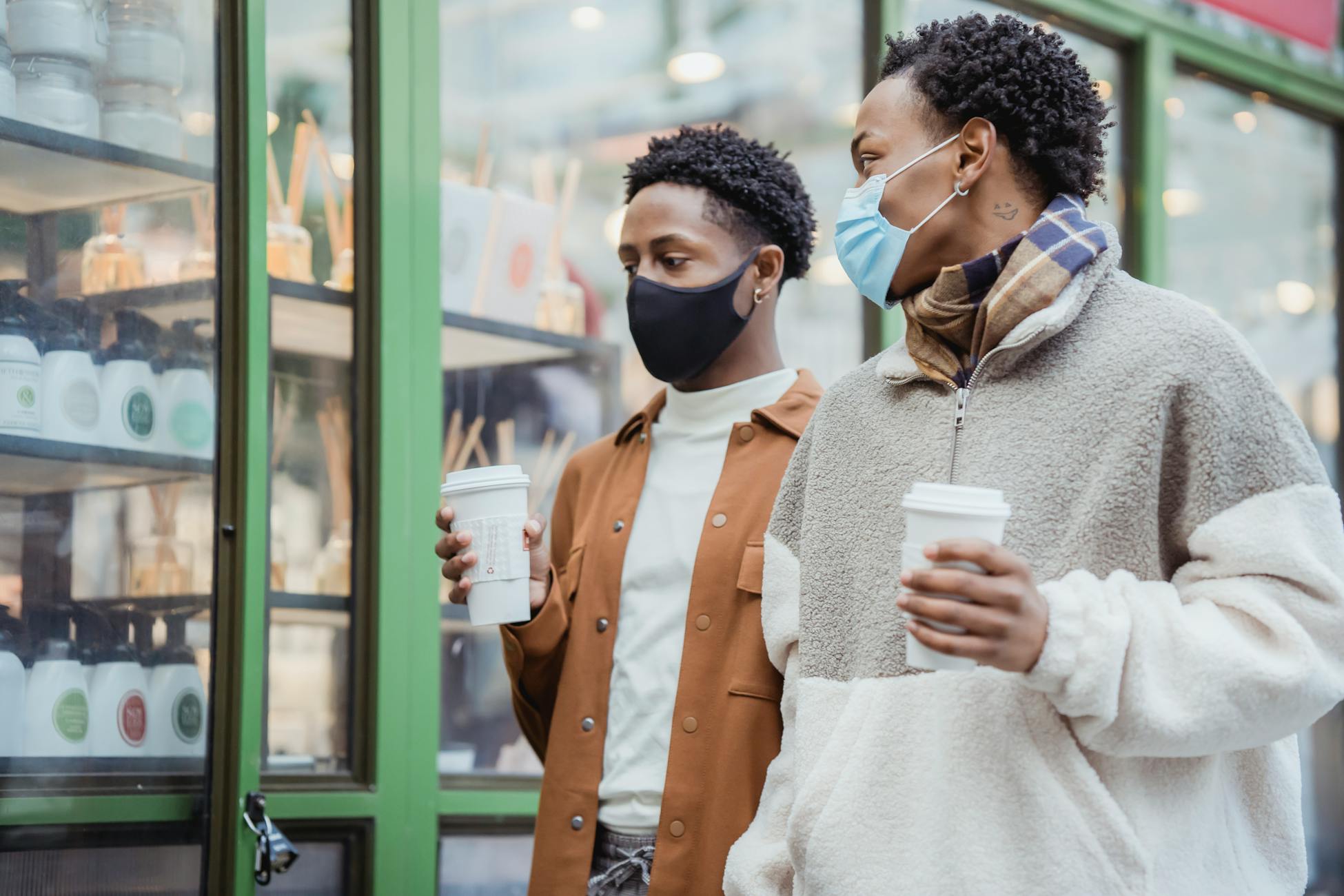 Stylish young African American male friends in warm clothes and medical masks walking on city street with coffee to go in hands and communicating
