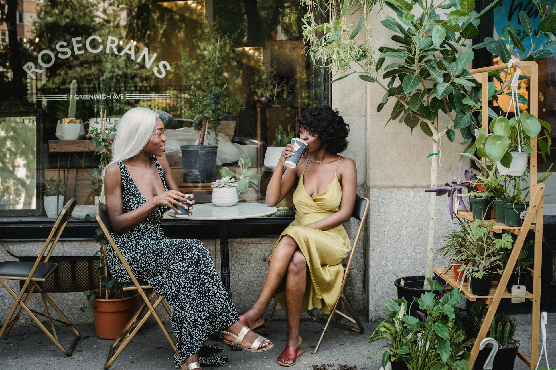 Two women sit at an outdoor café enjoying coffee surrounded by plants. Casual and relaxed atmosphere.
