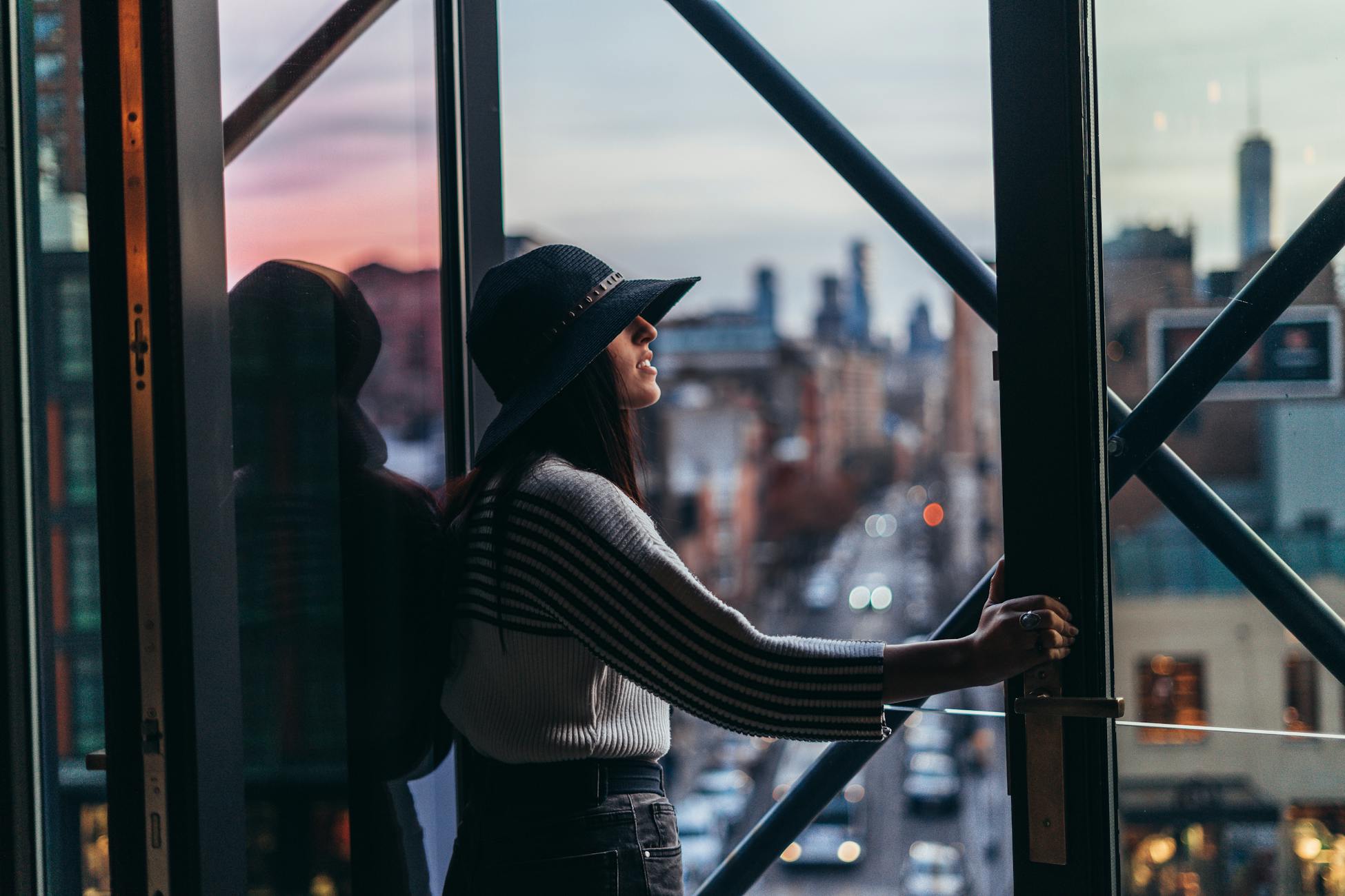 A woman wearing a blue hat stands by a window, gazing at the city skyline at dusk.