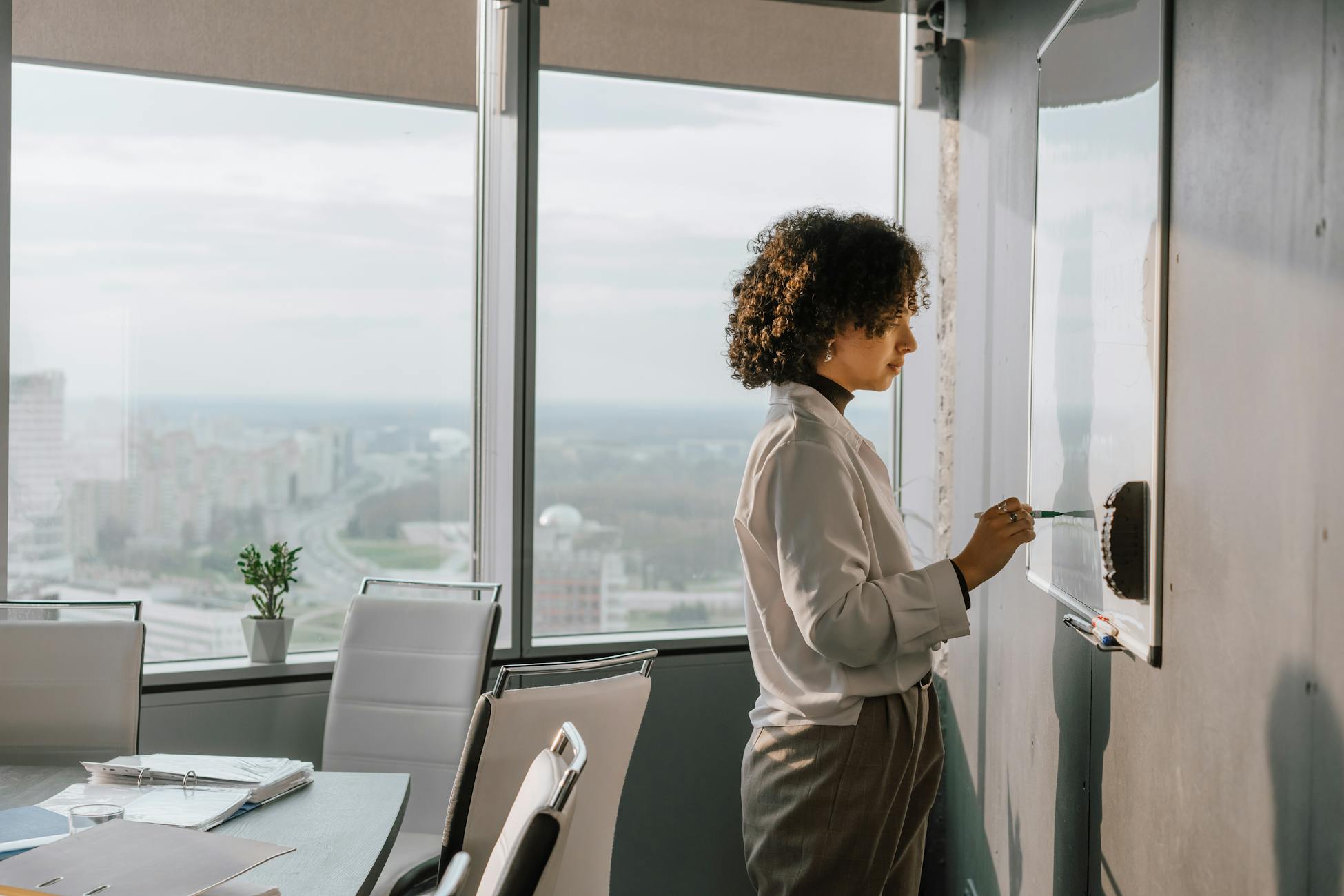 Confident woman with curly hair writes on a whiteboard in a modern office with city views.