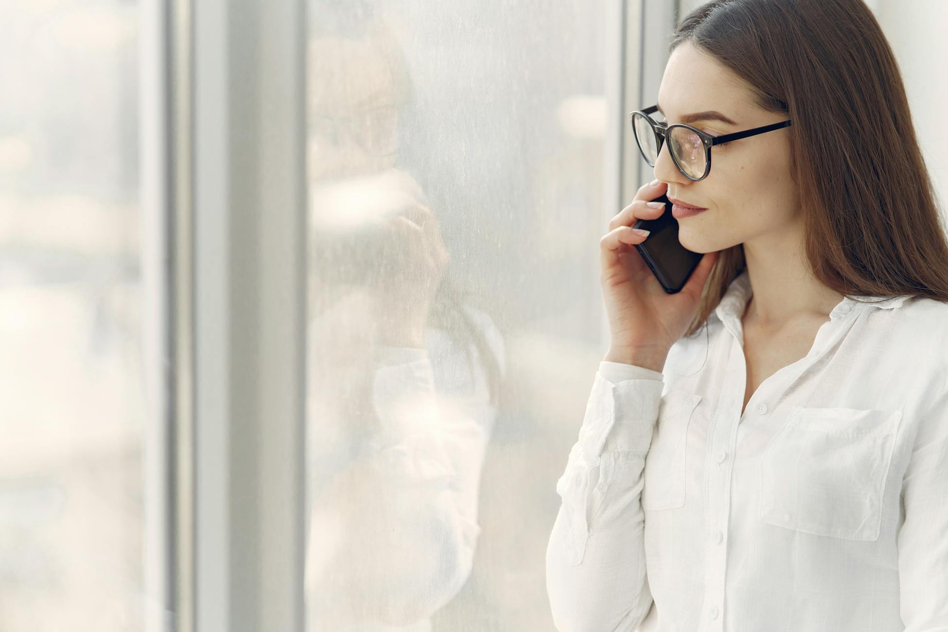 Pensive young woman in casual wear and eyeglasses speaking on mobile phone and looking at window while resting in modern apartment