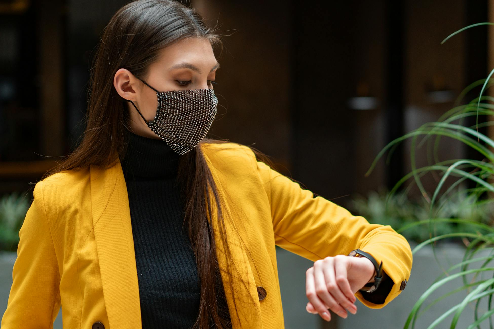 Fashionable woman in yellow blazer with a face mask checking her wristwatch in an indoor setting.