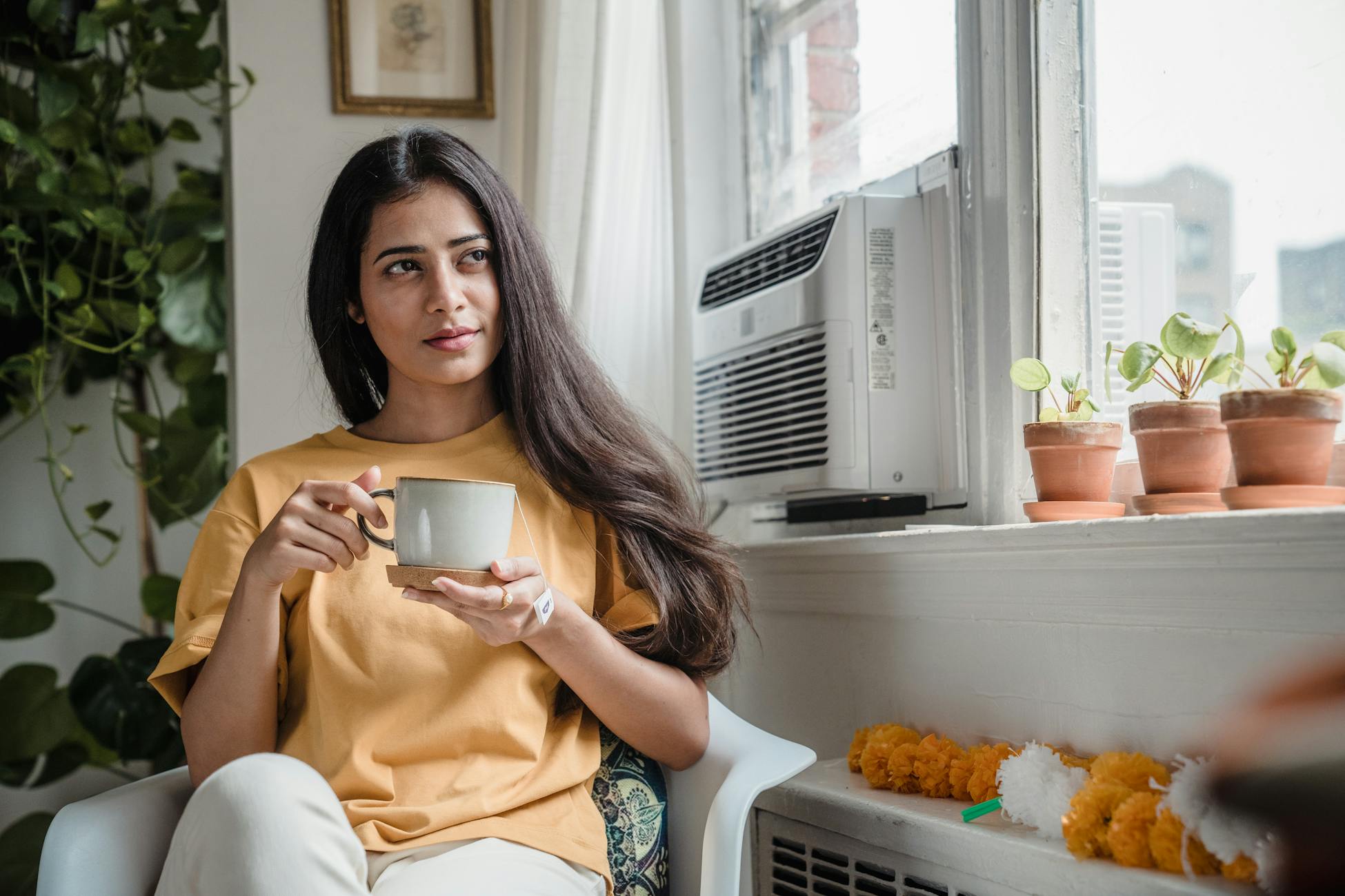 Indian woman sipping coffee indoors, surrounded by plants. A peaceful moment by the window.