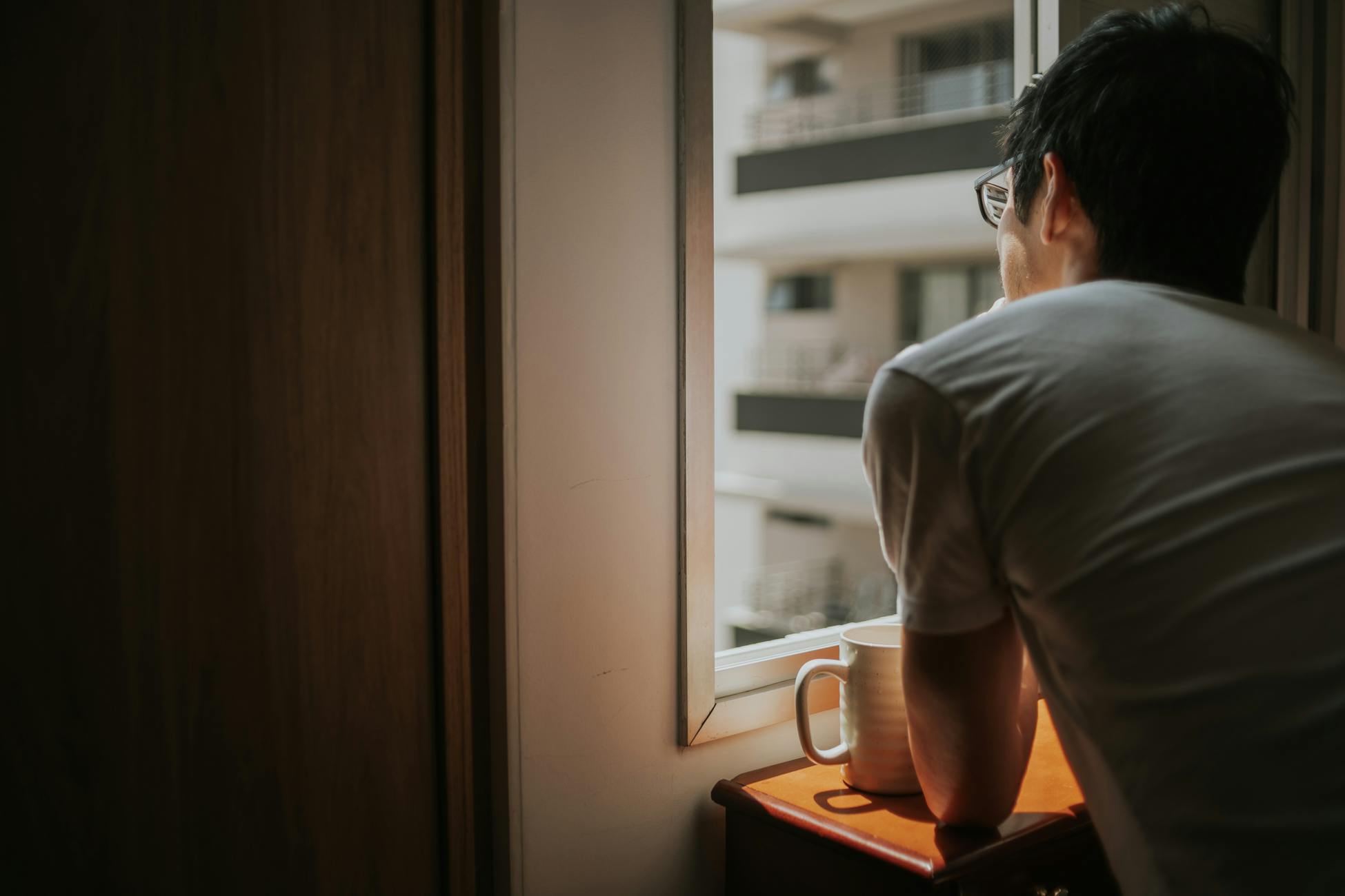 A man thoughtfully looks out a window with city buildings in view, holding a mug.