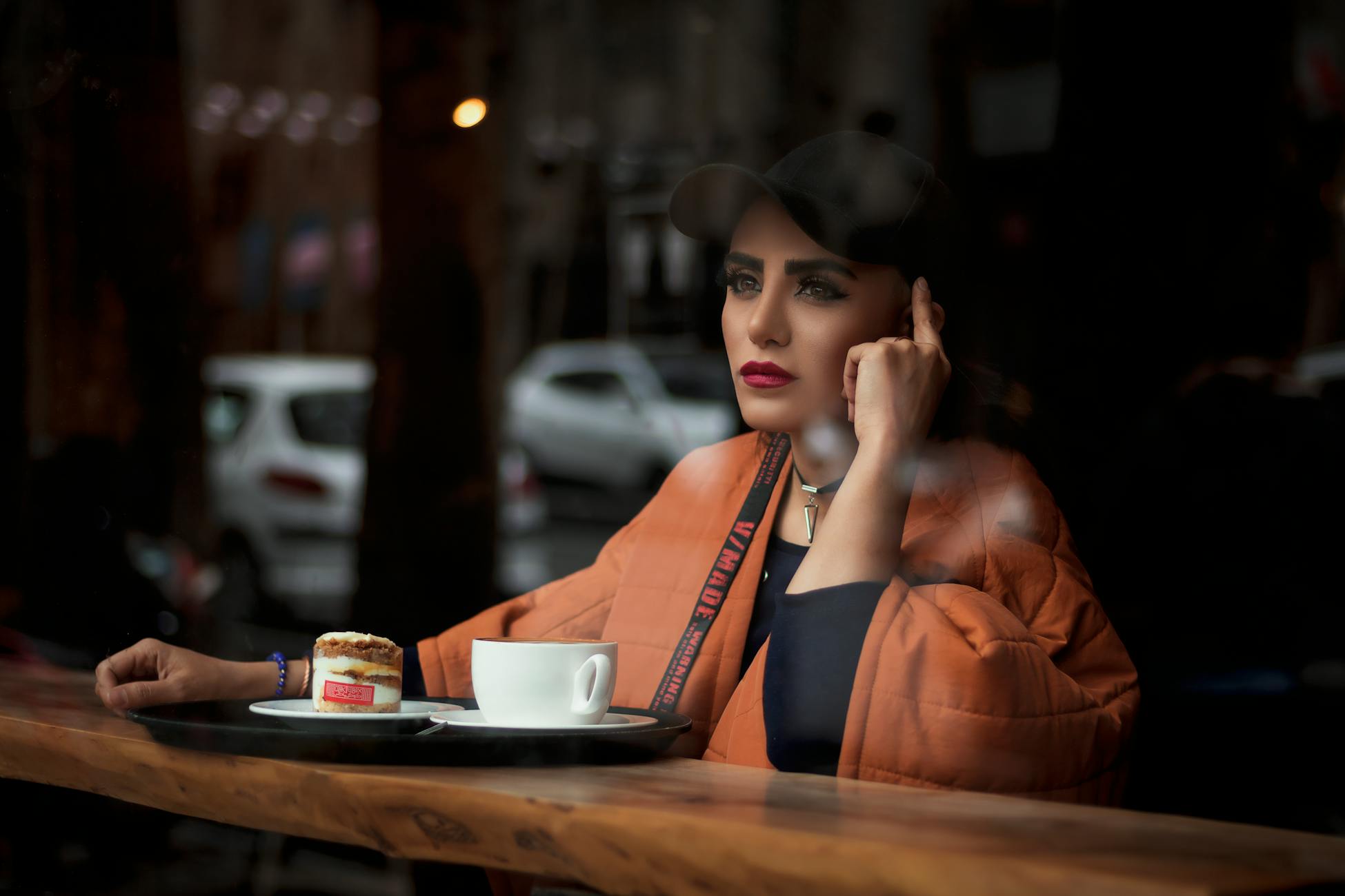 A woman in a cap and red lipstick sits in a Tehran café, reflecting over coffee and pastry.