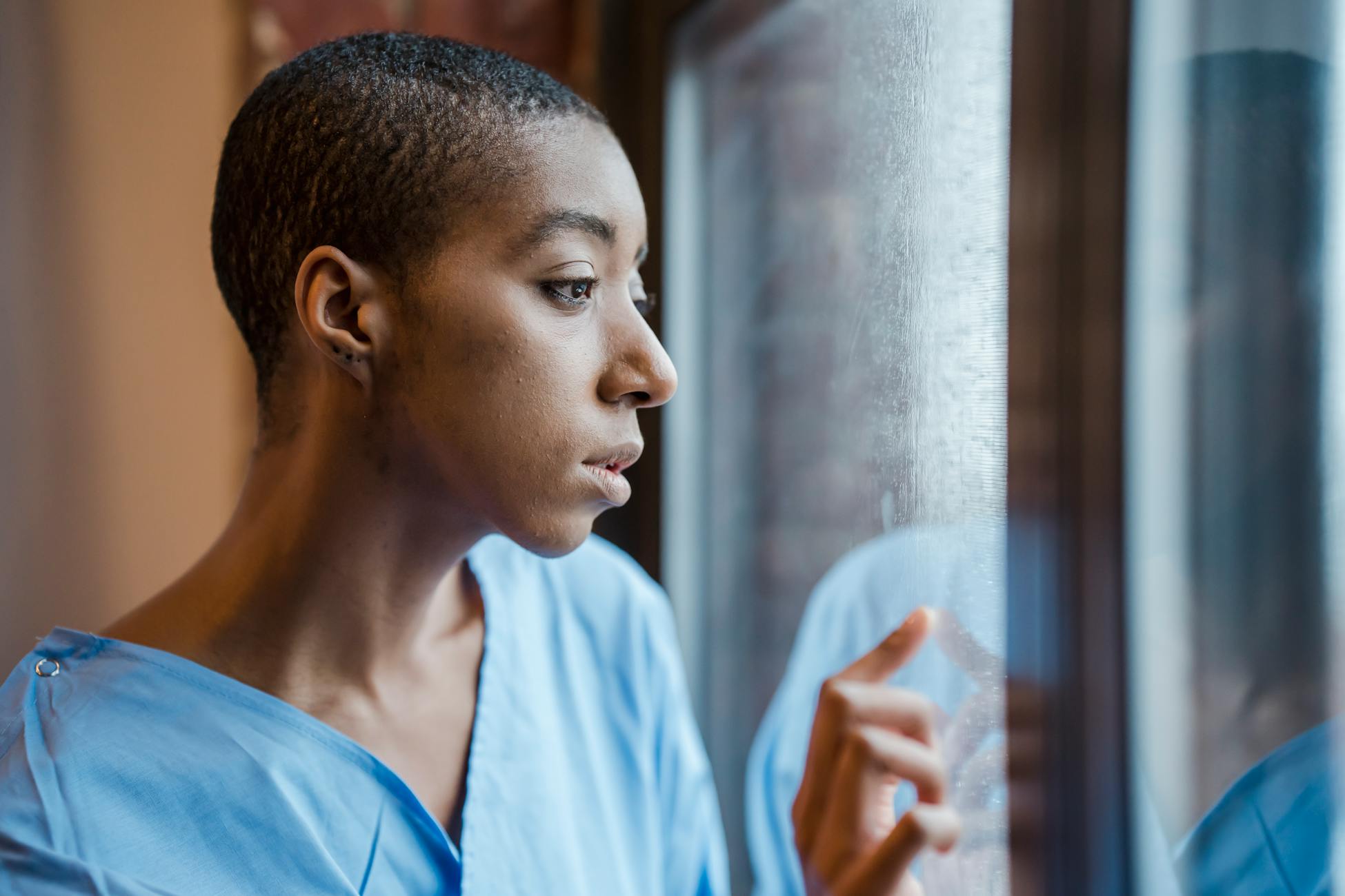 Upset African American female patient in medical wear touching window while looking away with concern