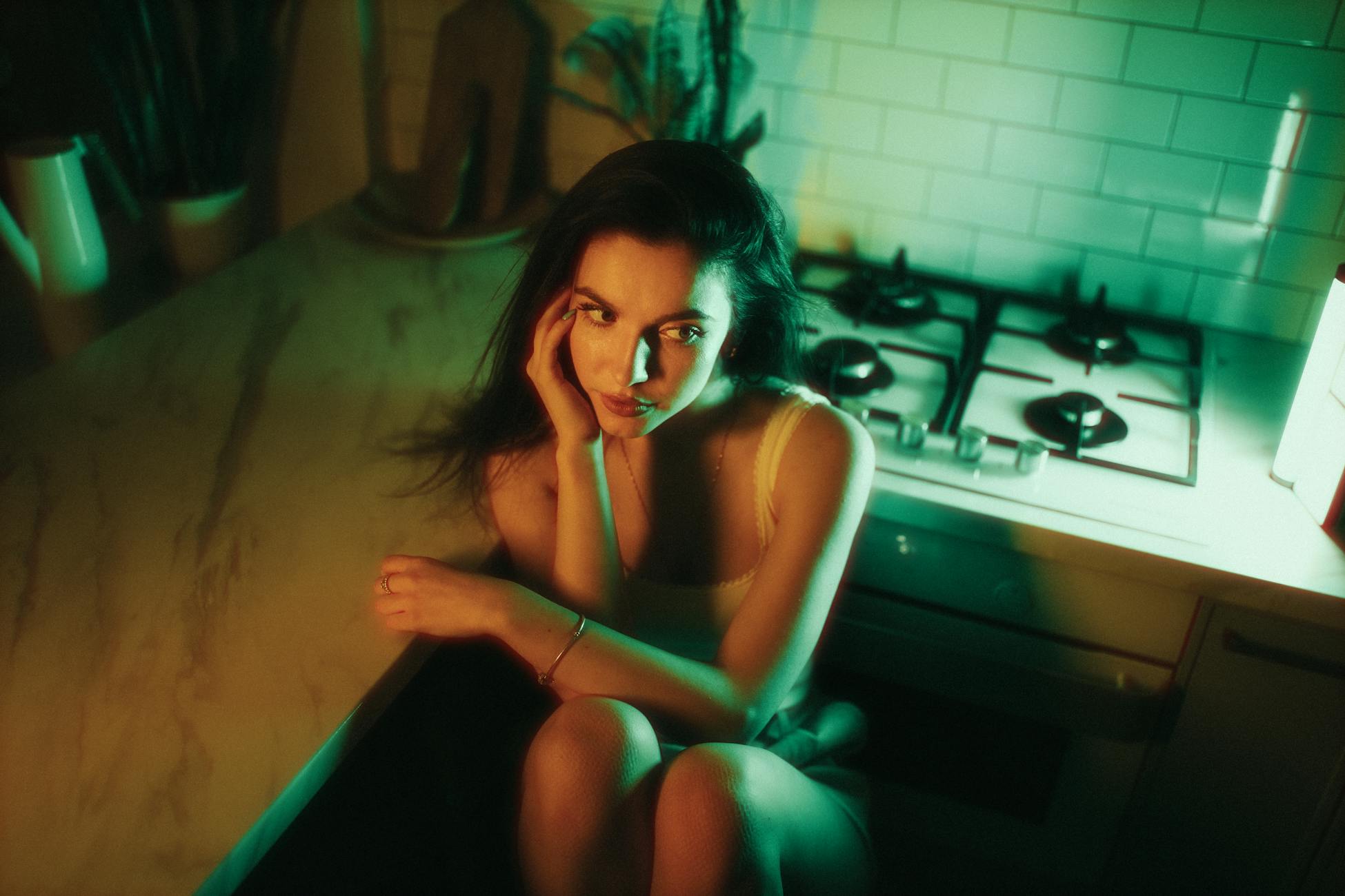A woman seated in a dimly lit kitchen with moody green and yellow lighting.