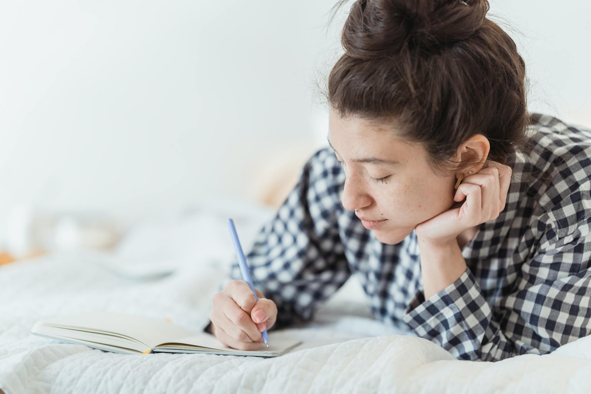 Young woman in checkered pajamas writing in a notebook while lying on a bed.