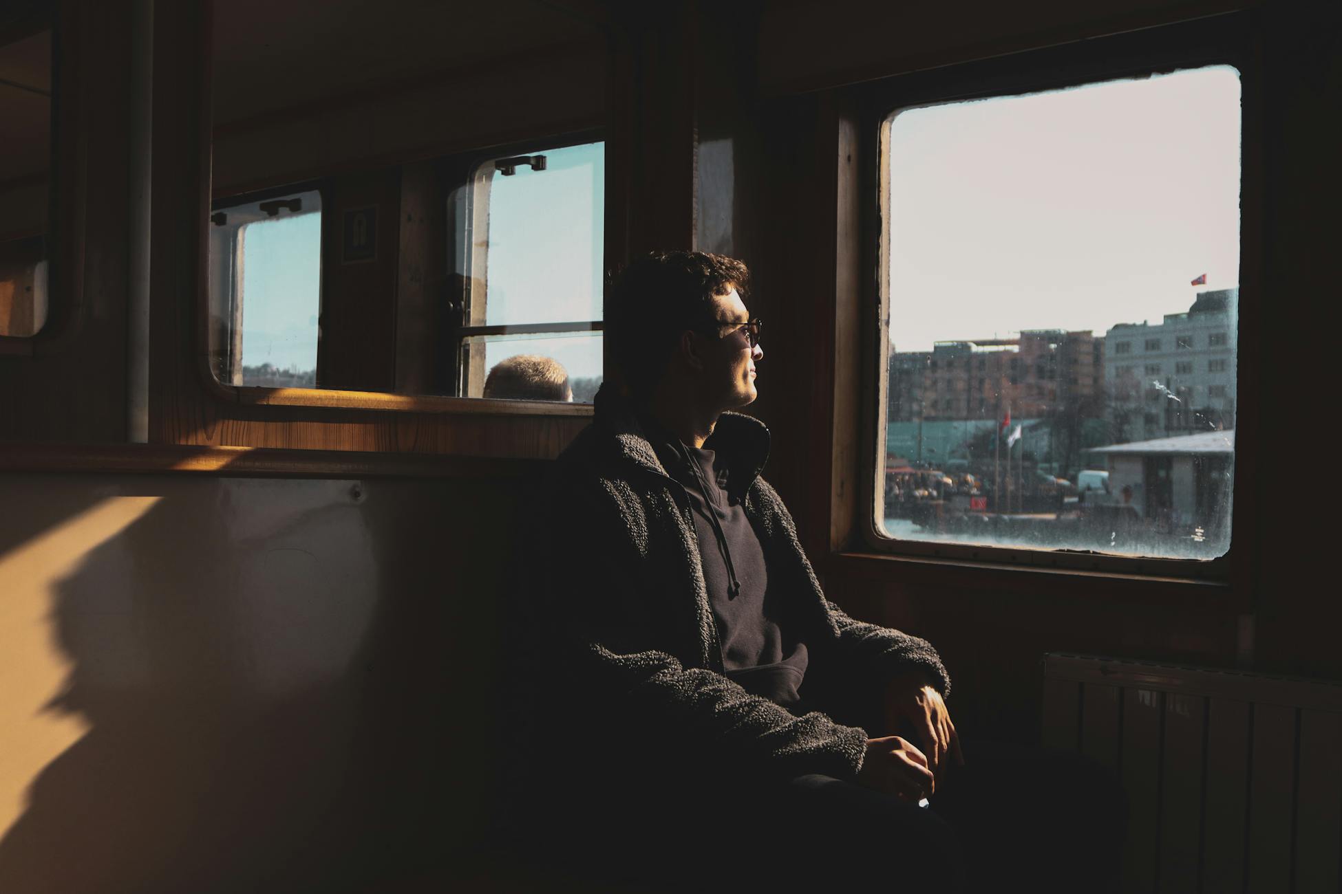 A man sits in contemplation, gazing out of a train window towards a cityscape in soft sunlight.