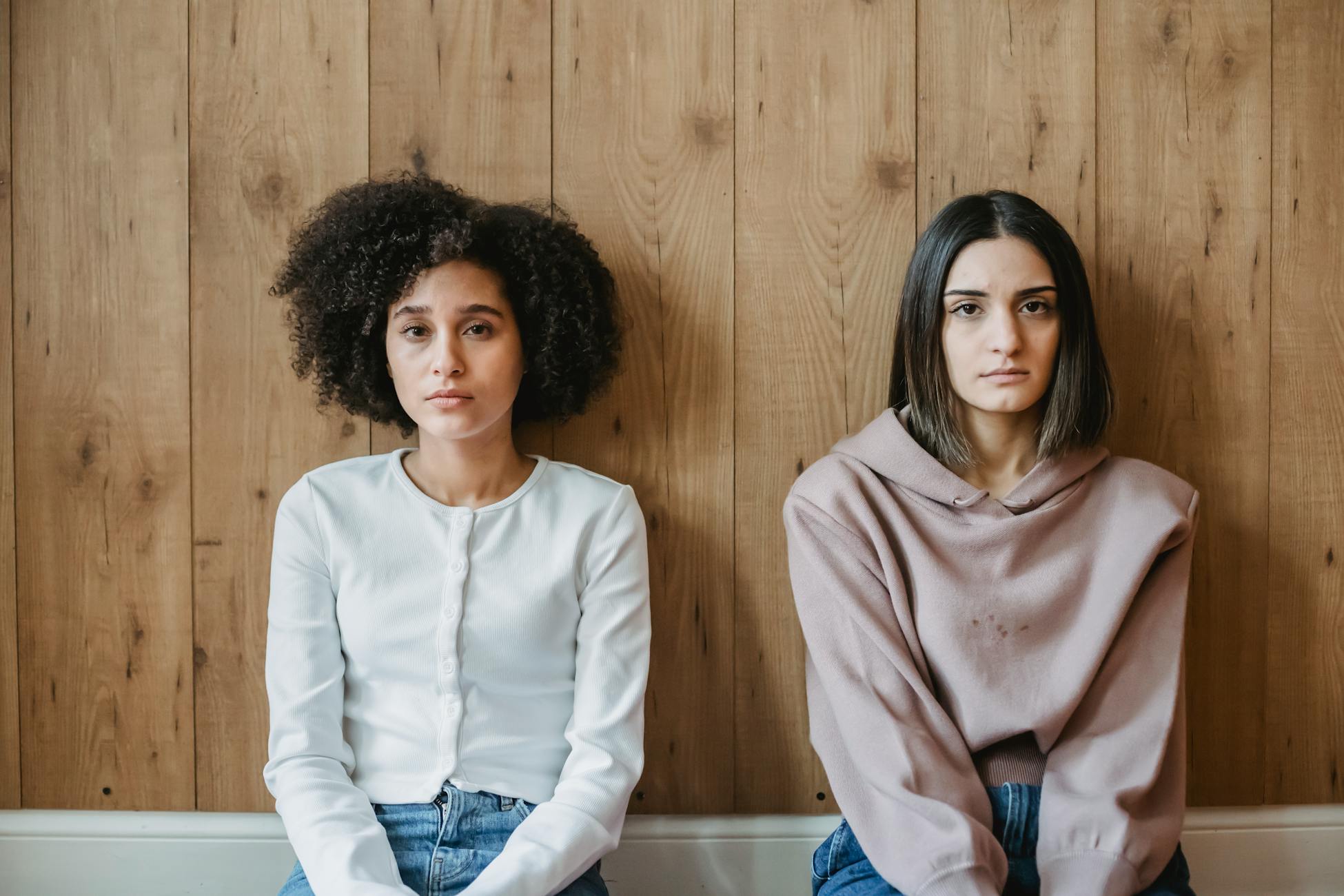 Pensive multiracial women in casual clothes sitting on floor against wooden wall and looking at camera in daytime
