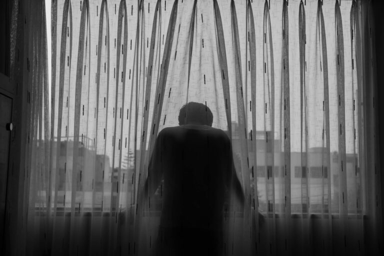 A black and white photo of a person standing behind a curtain, looking out the window, conveying isolation.
