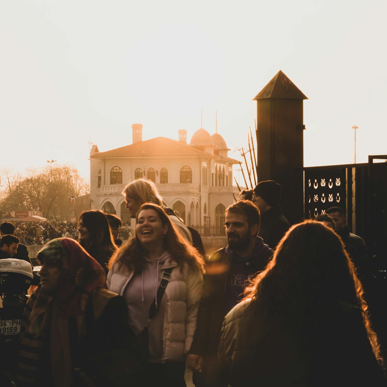 A lively crowd gathers near a historic building as the sun sets, casting warm light over the scene.