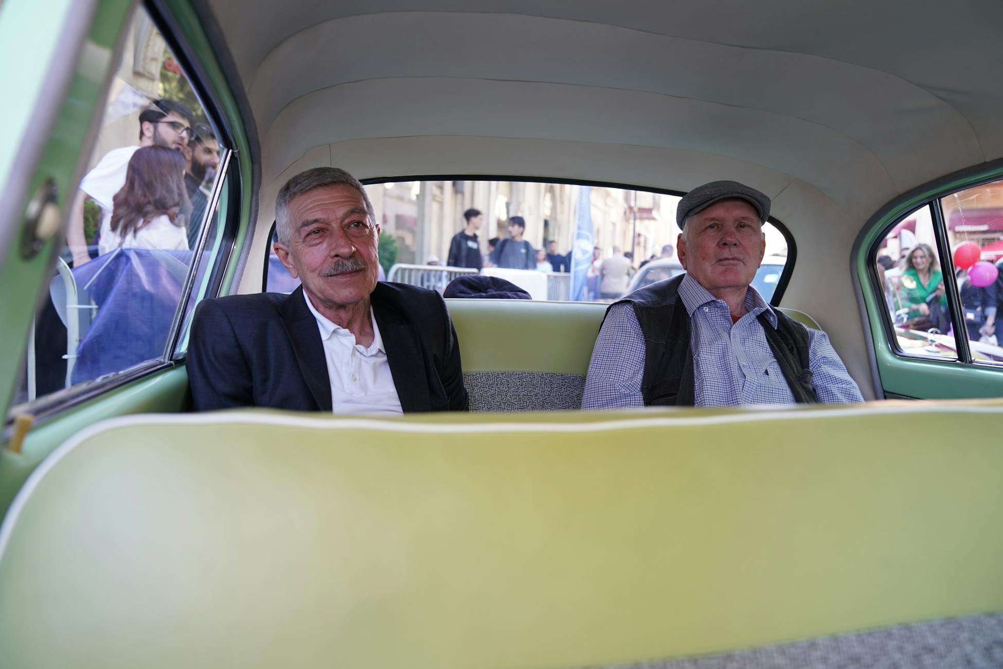 Two senior men relaxing in the back of a vintage car, enjoying a street scene.