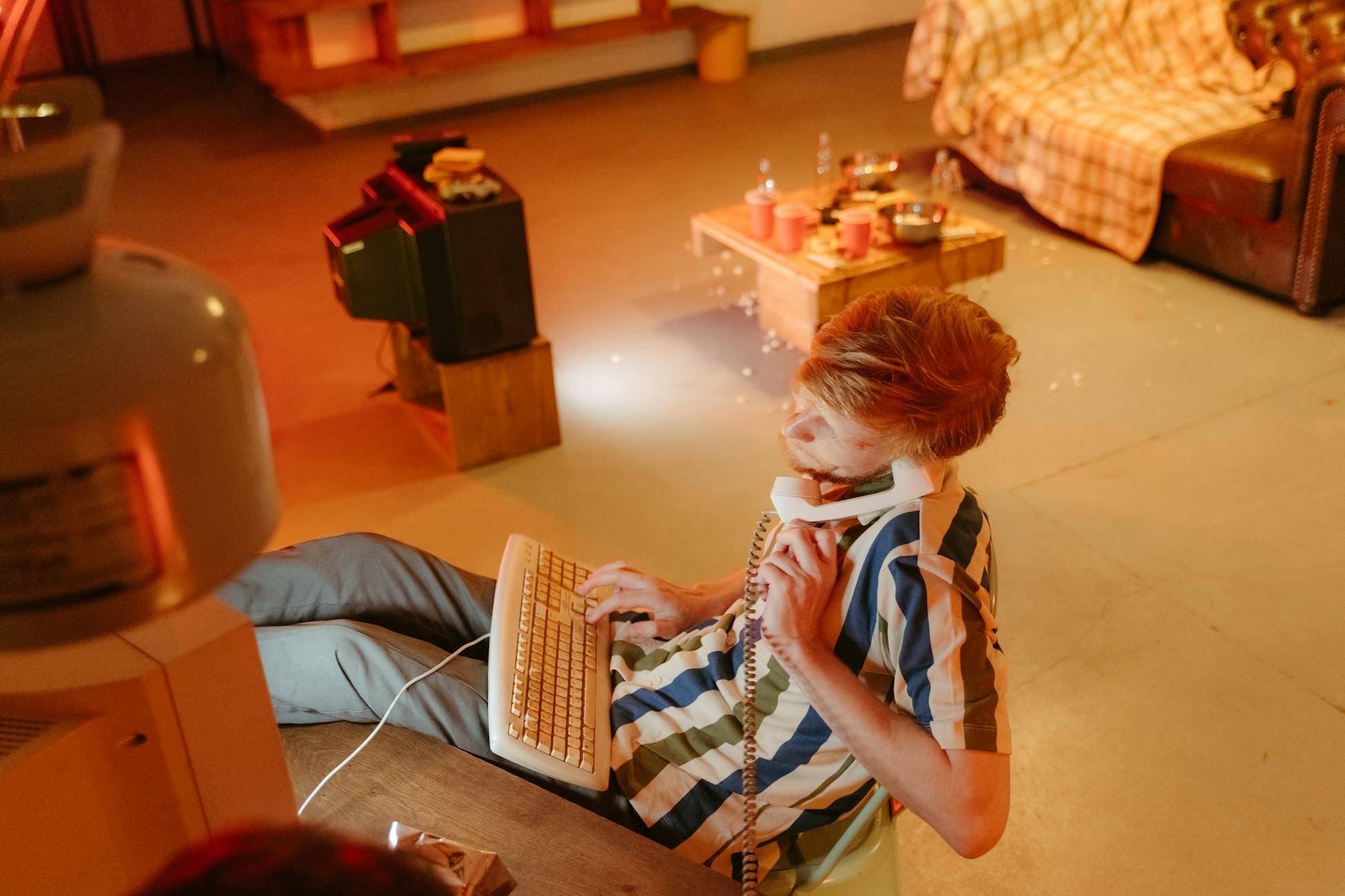 A man immersed in 80s tech, using a computer and phone in a retro-styled room.