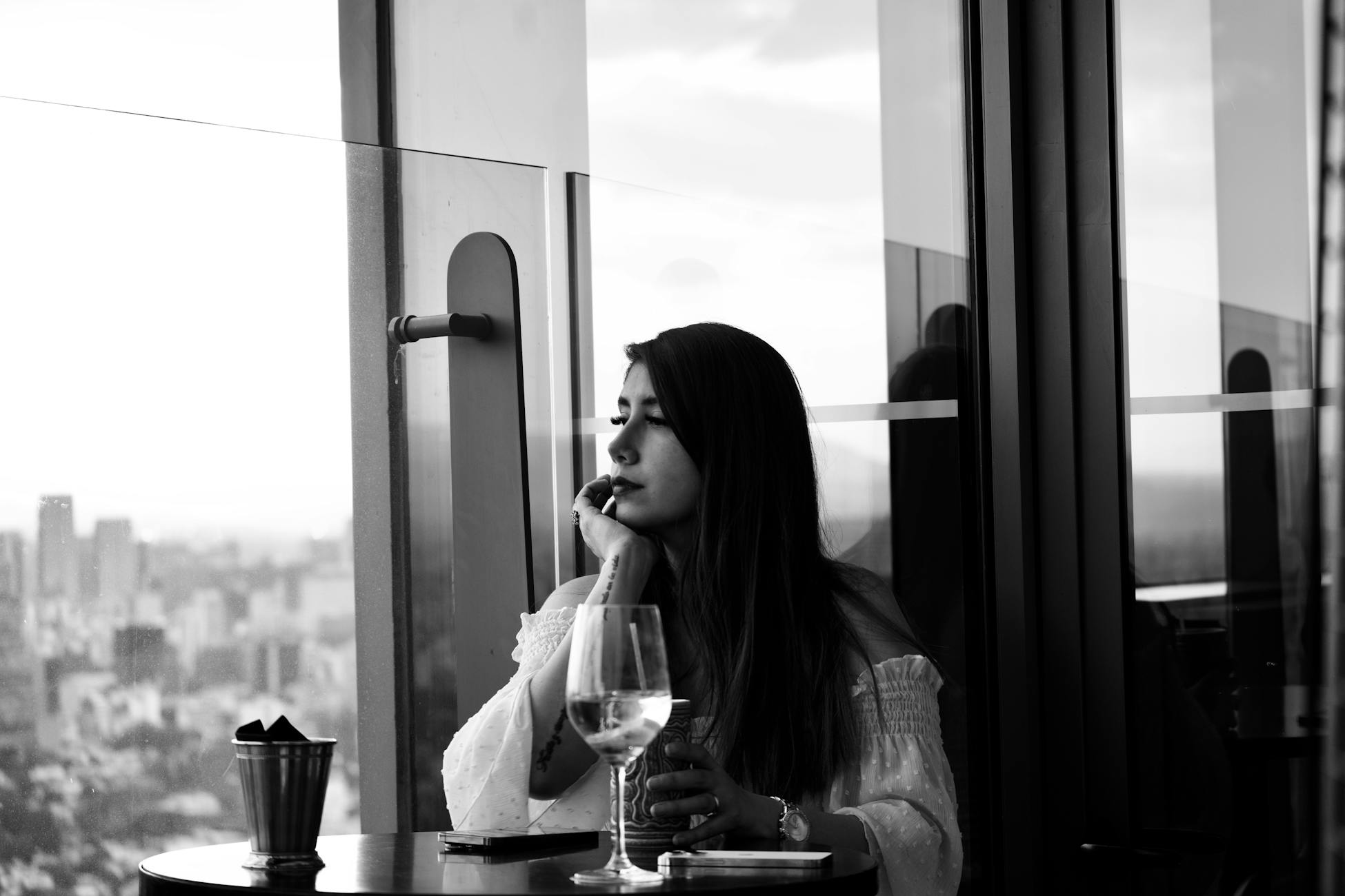 Black and white photo of a woman pondering at a rooftop bar with city views.