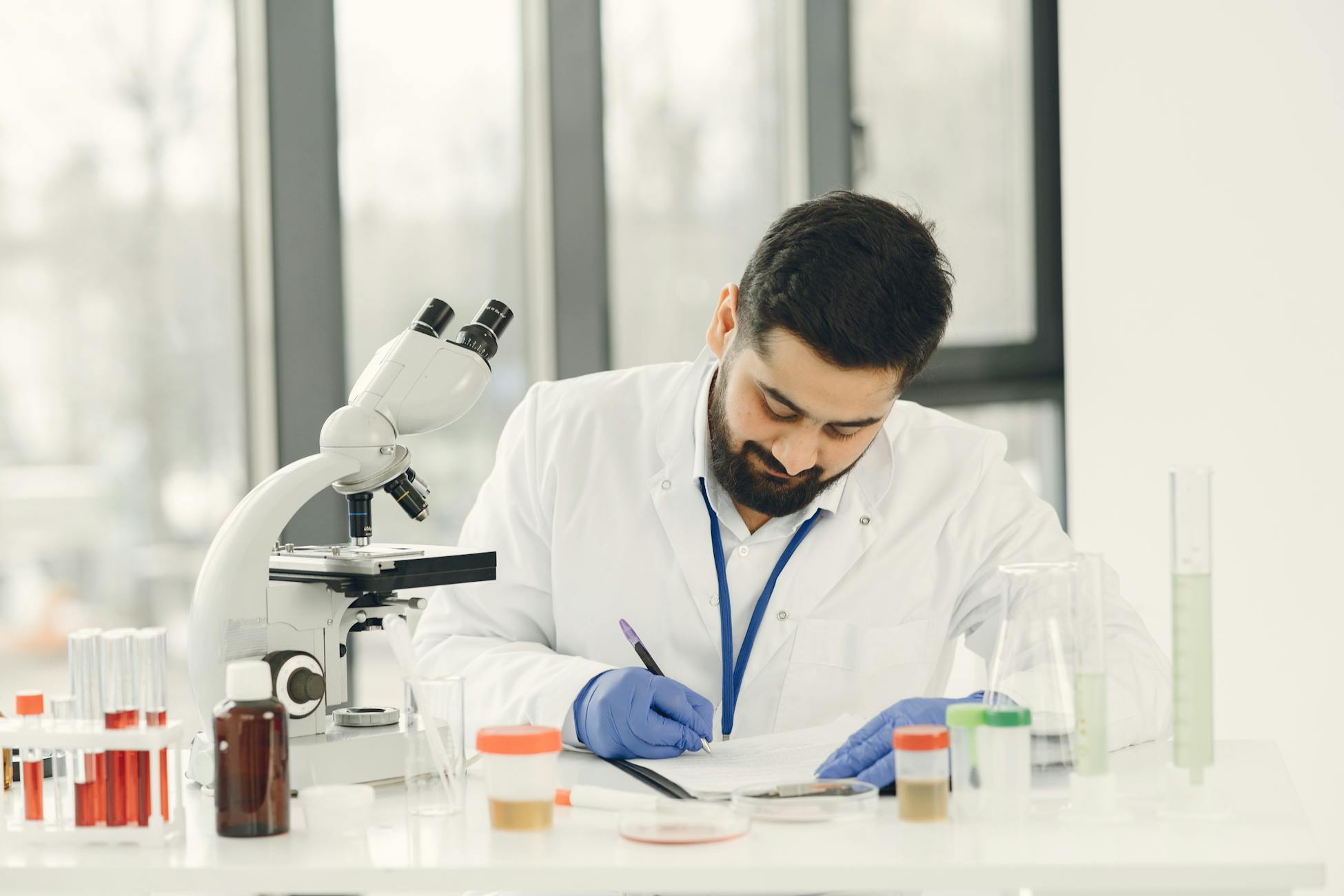 Scientist focused on writing notes amidst laboratory glassware and a microscope.