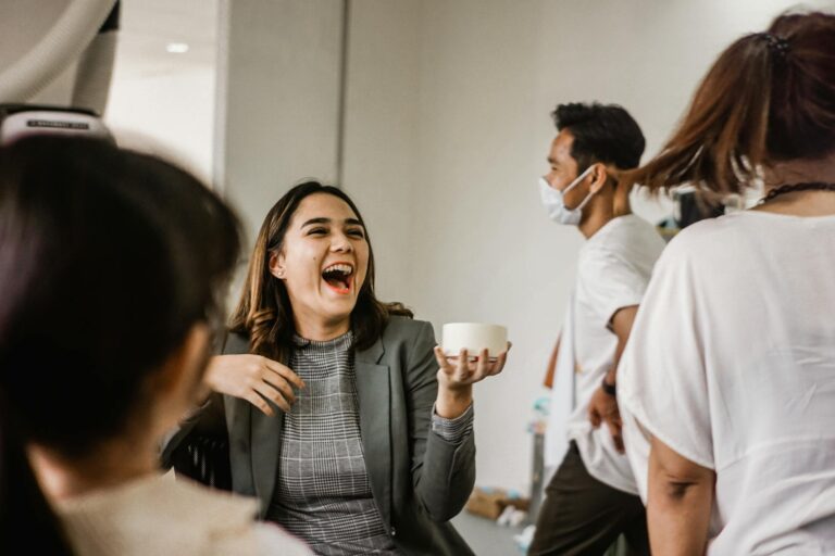 A woman in a gray blazer shares a laugh with colleagues in a lively office setting.