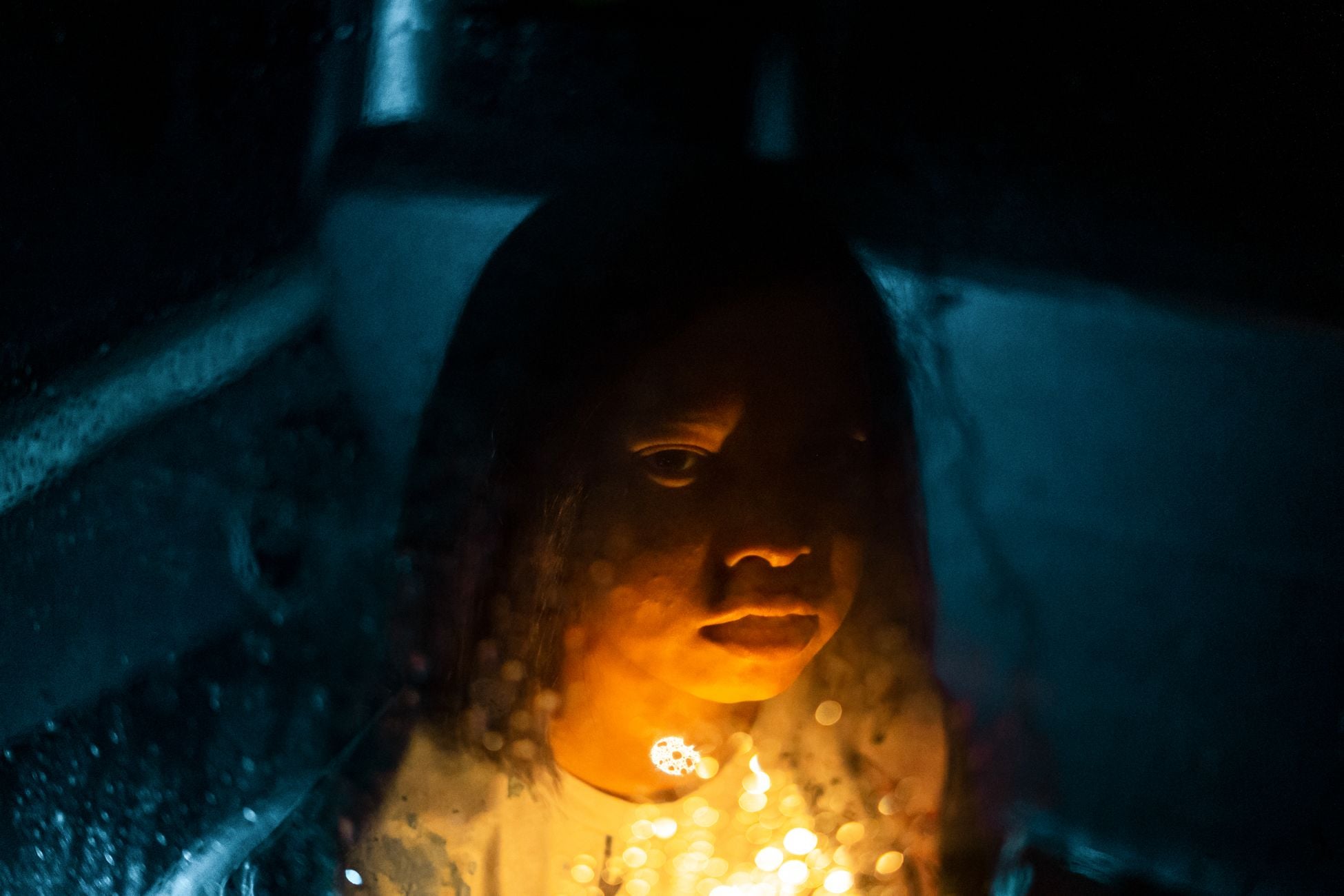 Through wet window view of thoughtful kid looking at camera with illuminated candle in dark room