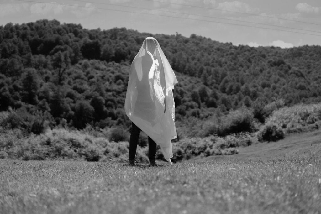 A solitary figure draped in white fabric stands in a serene meadow.