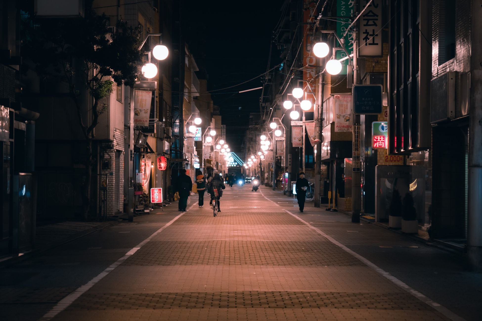 Dimly lit city street at night with glowing street lamps and few pedestrians.