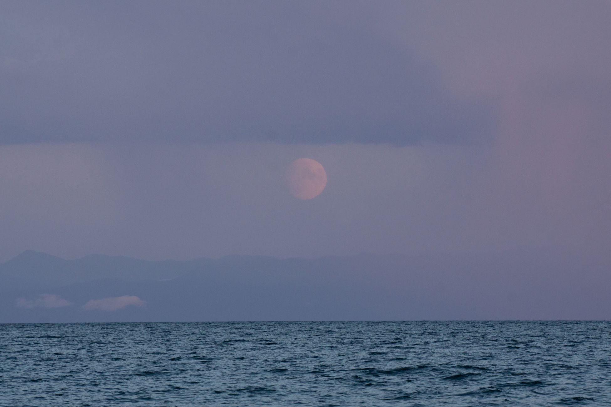 A serene view of the moon rising over a tranquil sea with a distant horizon at twilight.