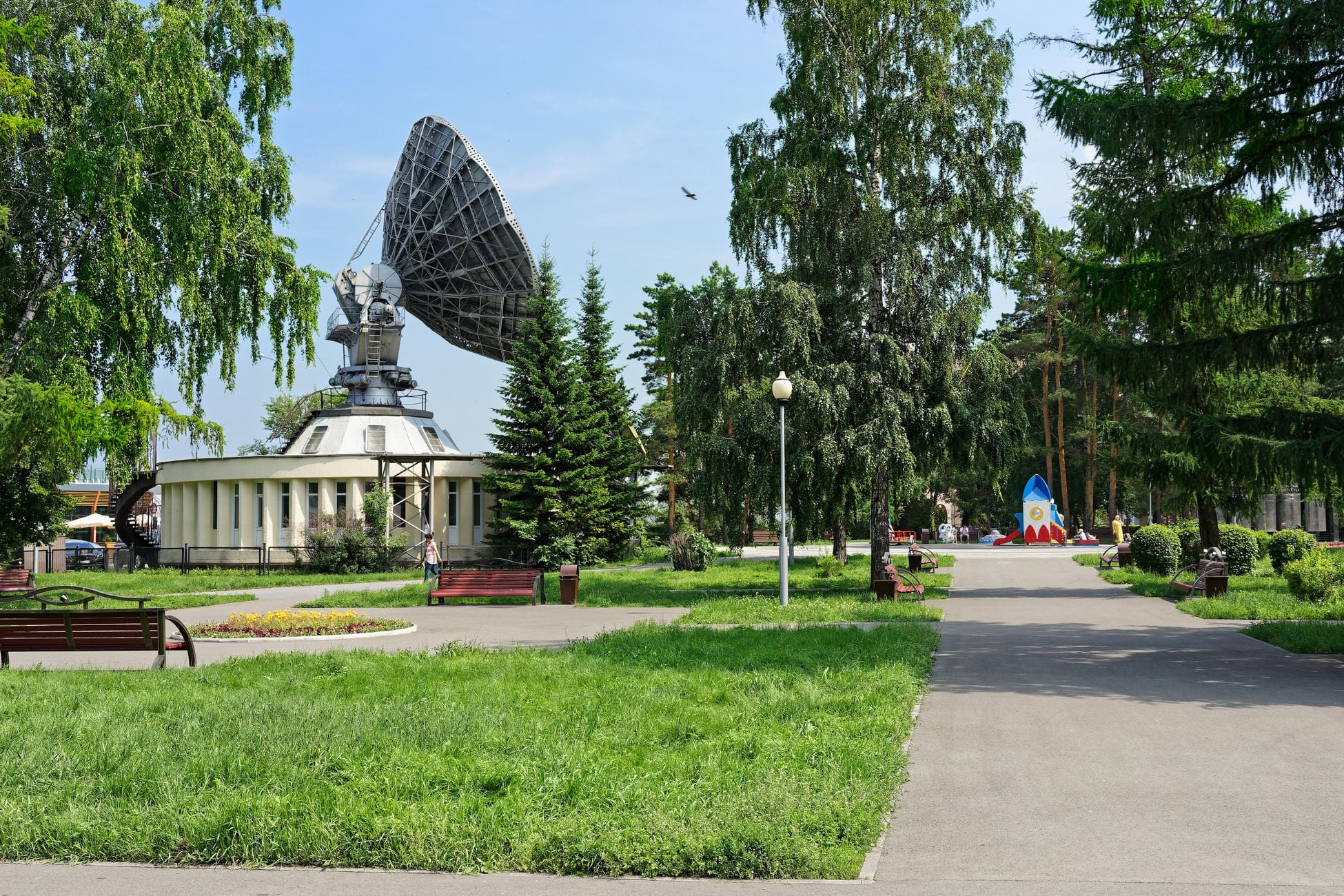 A serene park with pathways and a large satellite dish surrounded by lush greenery under a clear sky.