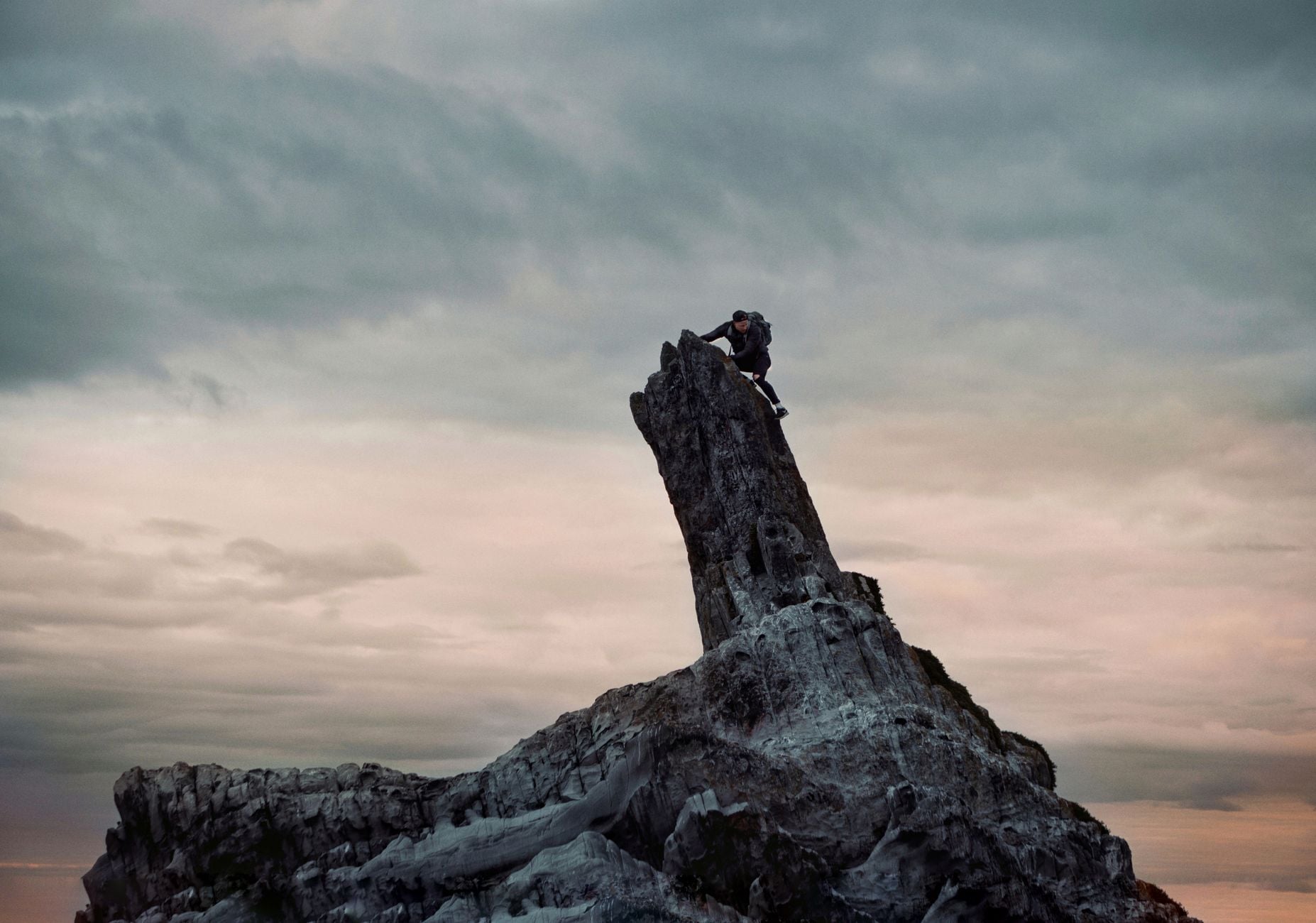 A climber scales a jagged rock formation under a dramatic cloudy sky at sunrise.