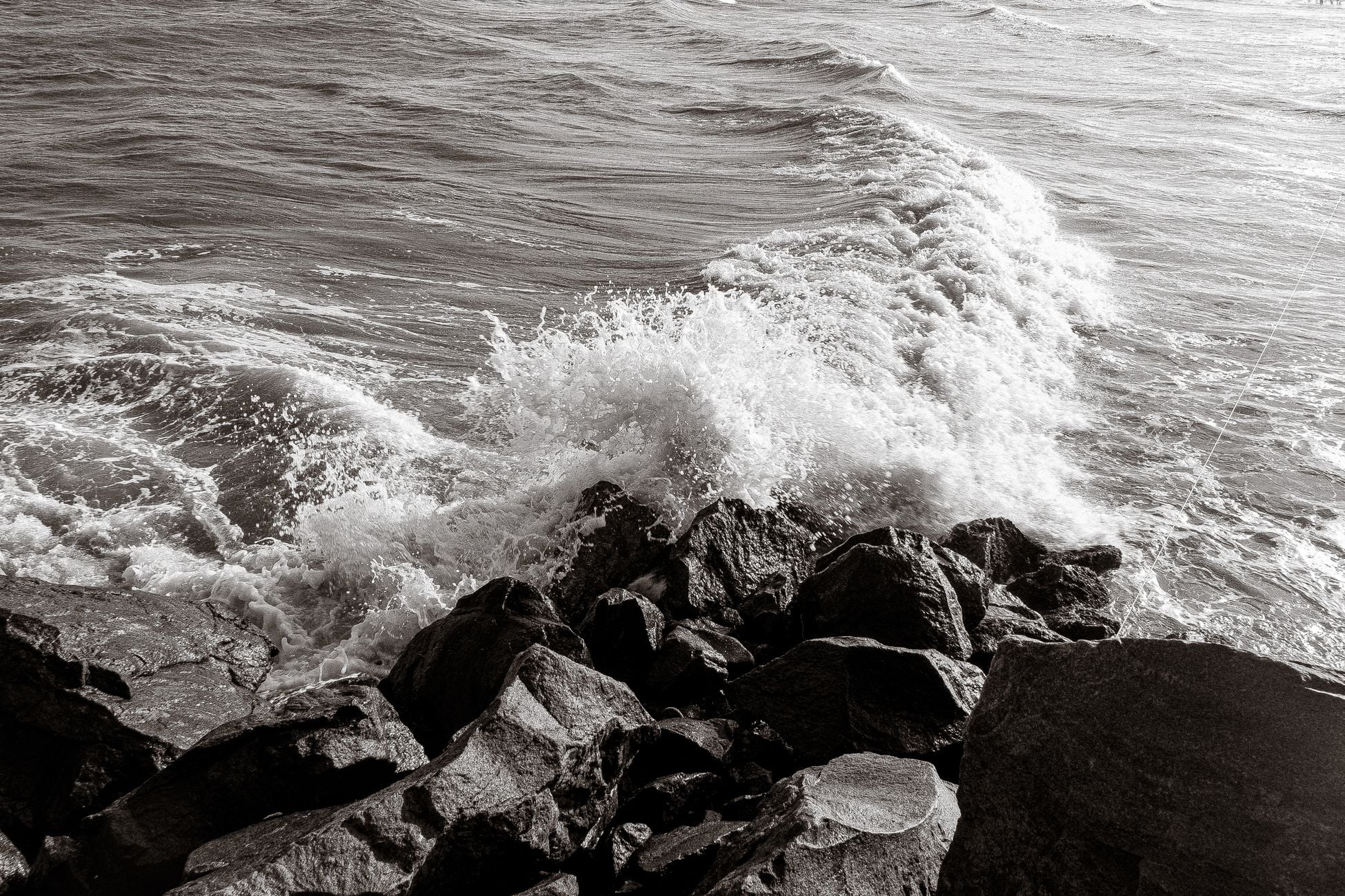 A powerful ocean wave crashes against a rugged rocky shoreline, captured in stunning black and white.