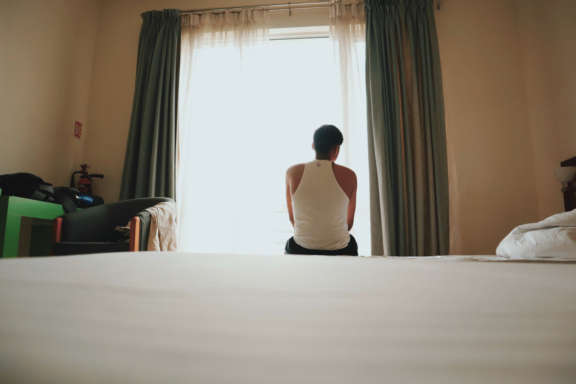 A man sits indoors facing a bright window, lost in thought, casting an introspective mood.