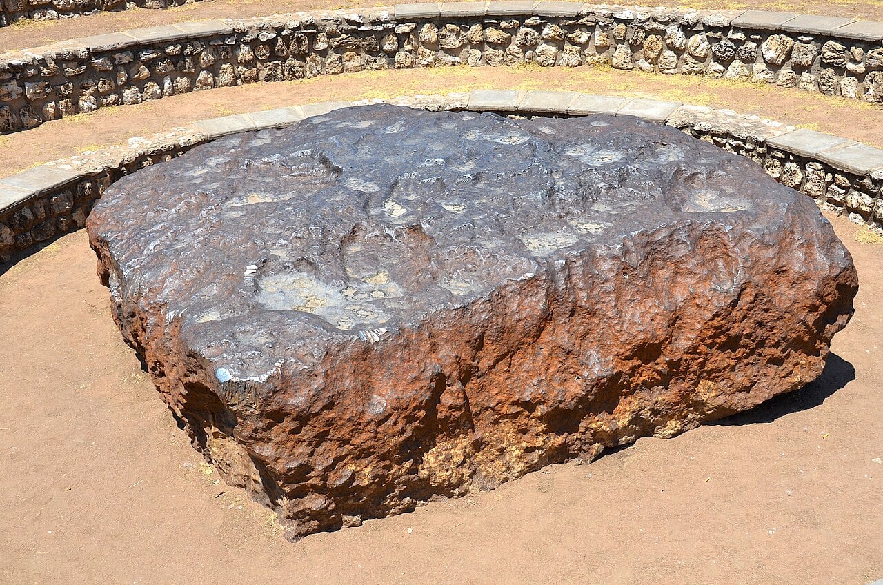 In 1920, a farmer ploughing near Grootfontein in Namibia struck a buried slab of iron weighing roughly 60 tonnes. It proved to be Hoba, the largest intact meteorite ever found, and it has never been moved from where it landed—probably less than 80,000 years ago. No crater survives around it; its broad, flat shape may have slowed its descent until it struck more like a colossal dropped anvil than a cosmic projectile.