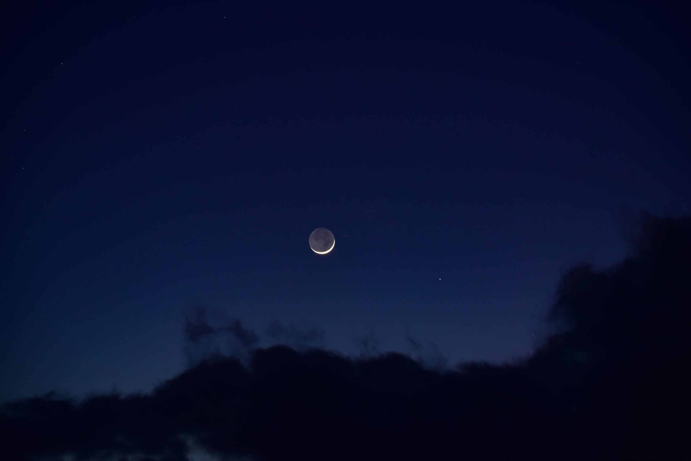 Young crescent Moon with faint earthshine in a deep blue sky above dark clouds