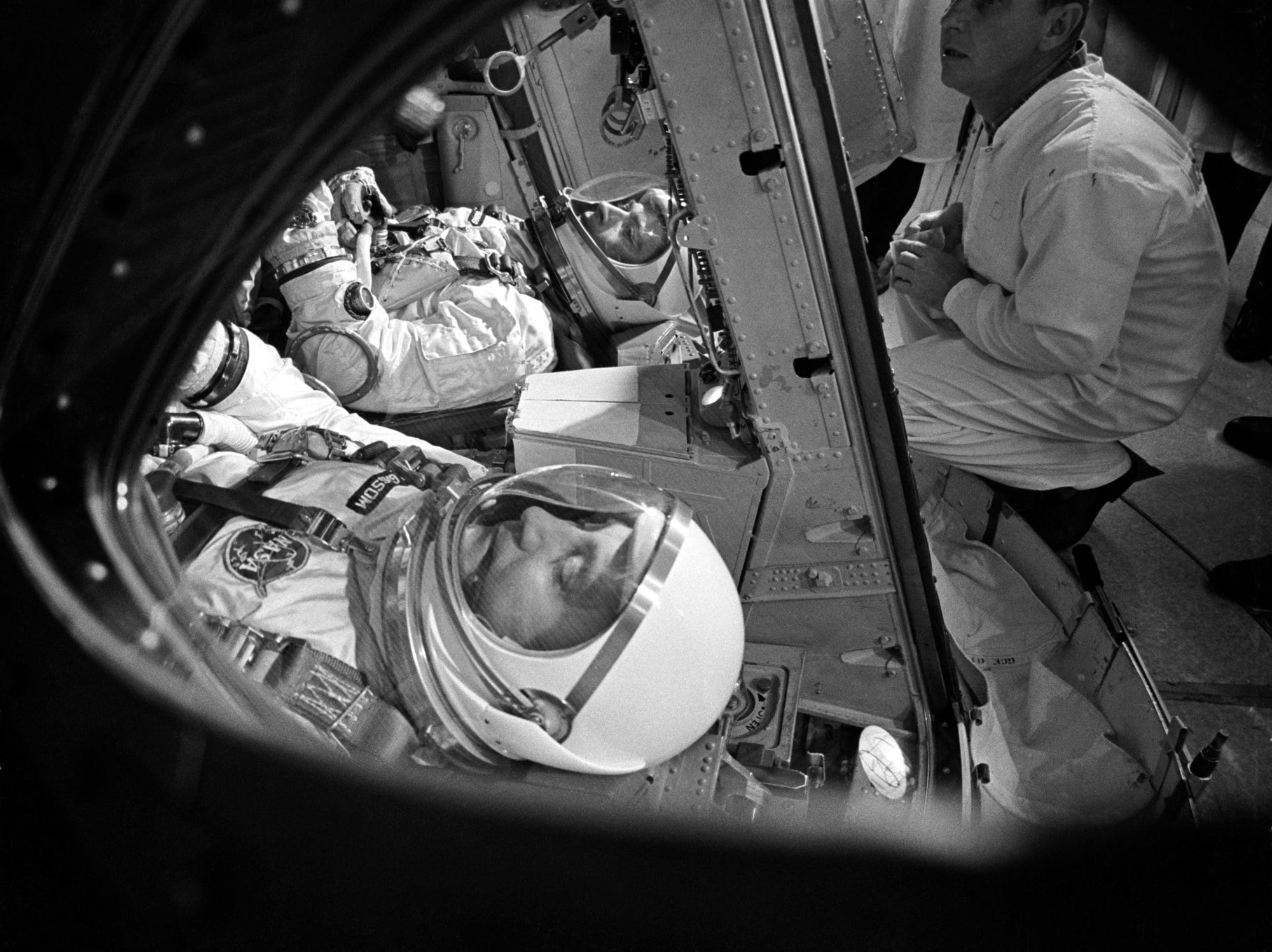Gus Grissom and John Young in spacesuits and helmets strapped into the Gemini III spacecraft, seen through the open hatch